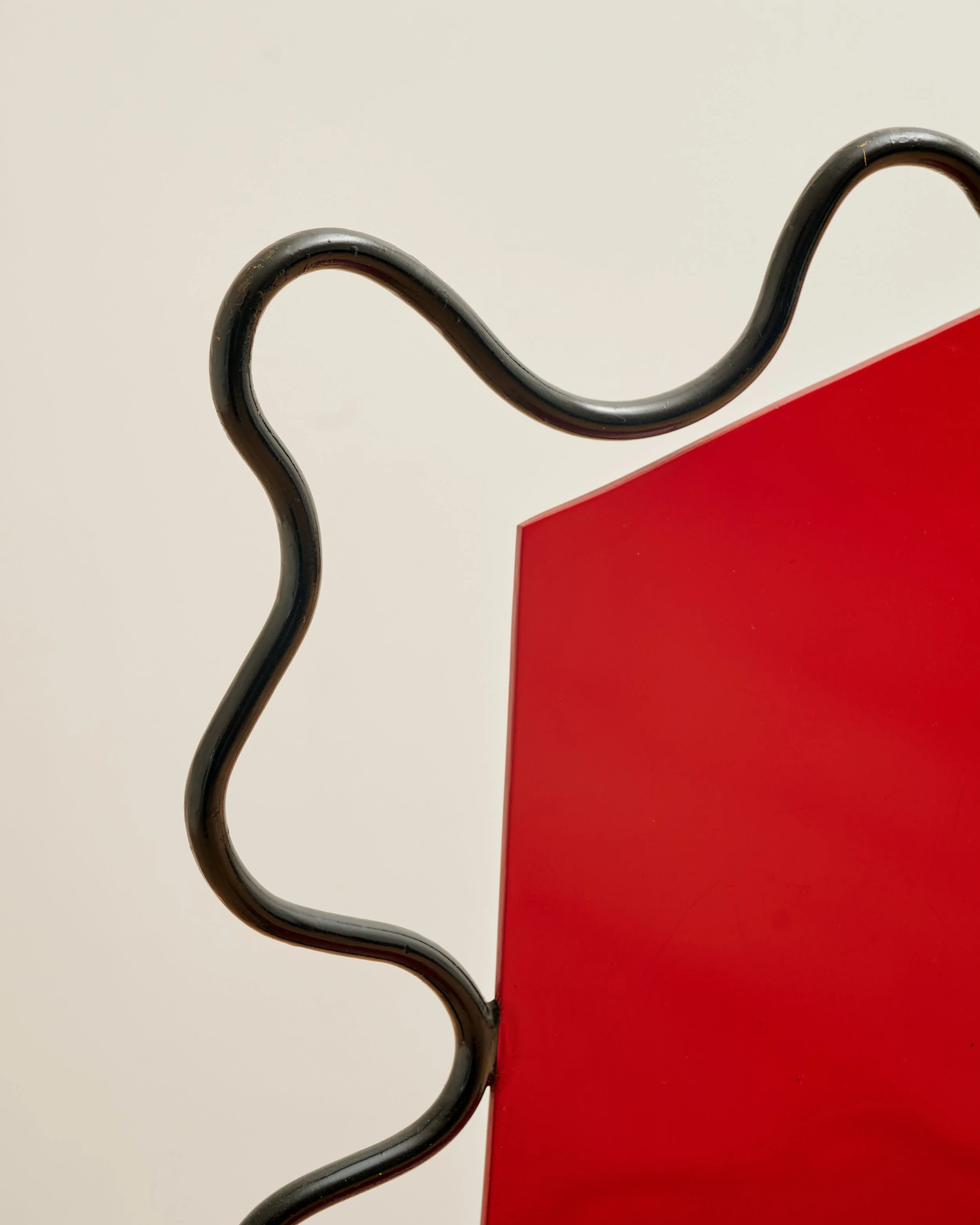 Close-up of a black metal hook attached to a red surface, with a plain off-white background. Howard Meister.