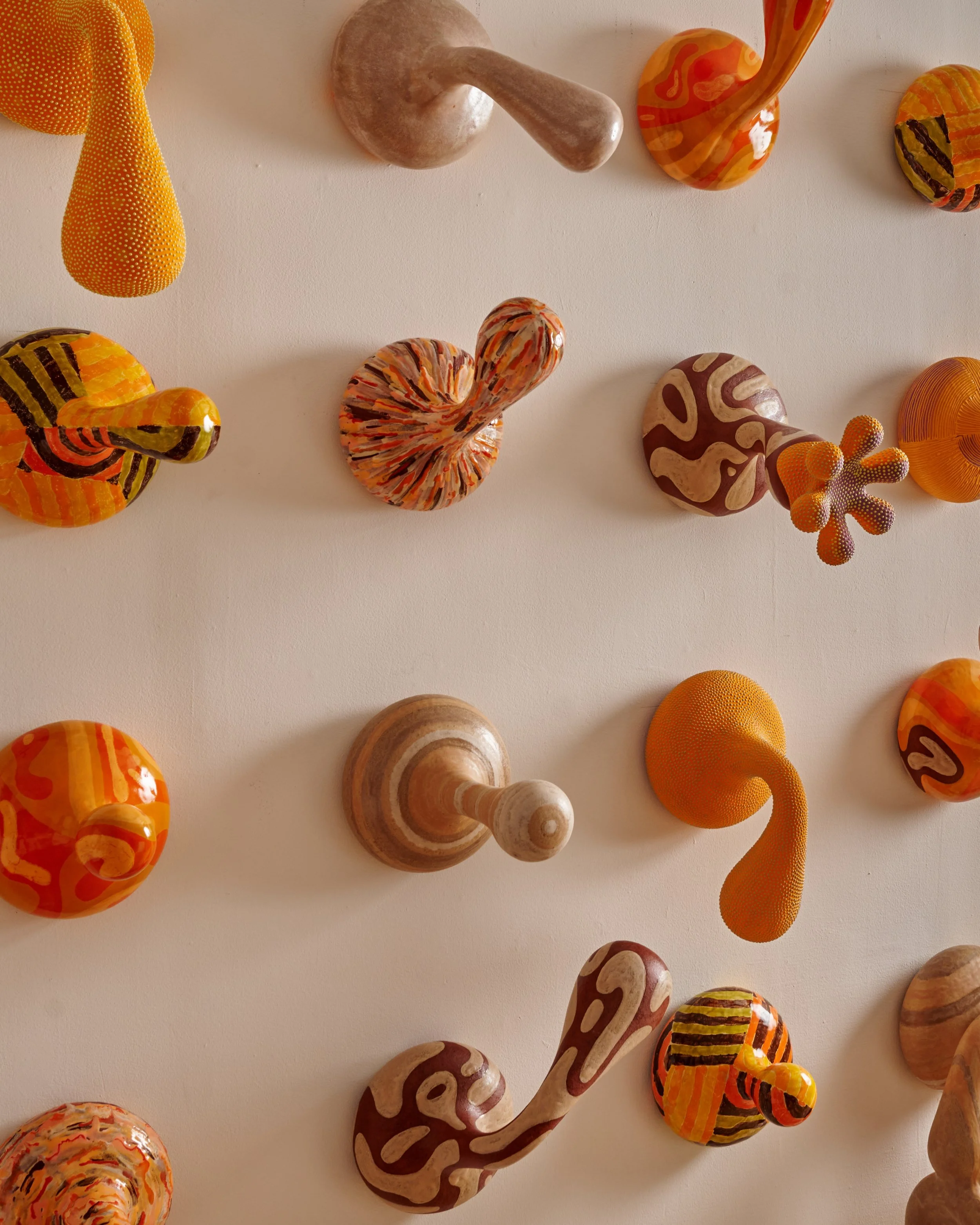 Colorful glass sculptures on a white surface. The sculptures vary in shapes, including swirls, spheres, and abstract forms, with a mix of warm tones like orange, yellow, and brown, some with striped or dotted patterns. Joël Brodovsky-Adams.