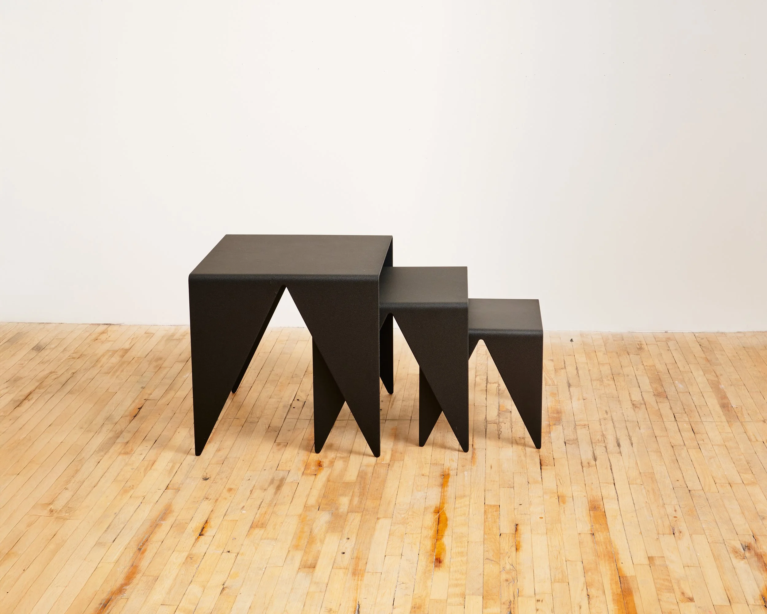 Three black nesting tables with angular legs, arranged on a wooden floor against a white wall.