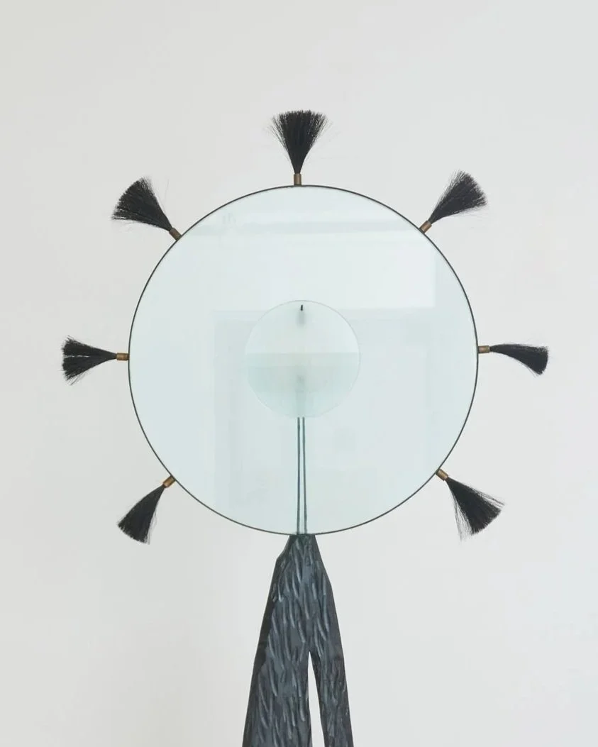 Modern wall clock designed to resemble a person with broomstick arms and head, featuring a round mirror face and tassel details. Alex Locadia.