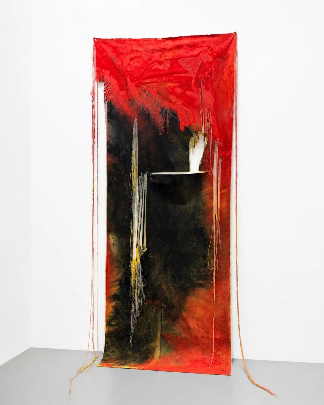 Abstract textile art piece with red, black, and white colors and hanging yarns, displayed on a white gallery wall. Vaughn Davis Jr.