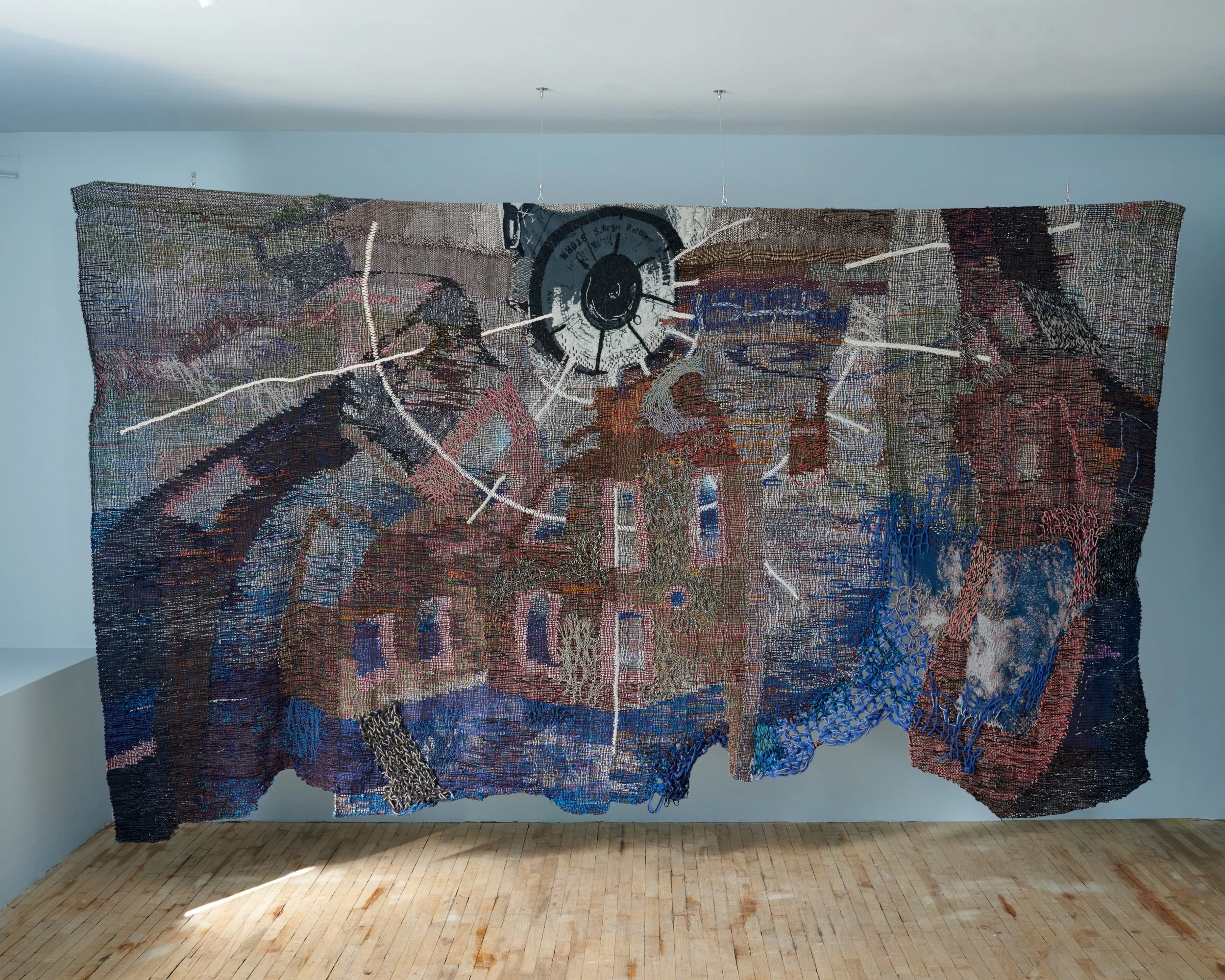 A large textile artwork hanging on a wall, featuring abstract patterns and a central clock-like design, with a range of colors and textures.
