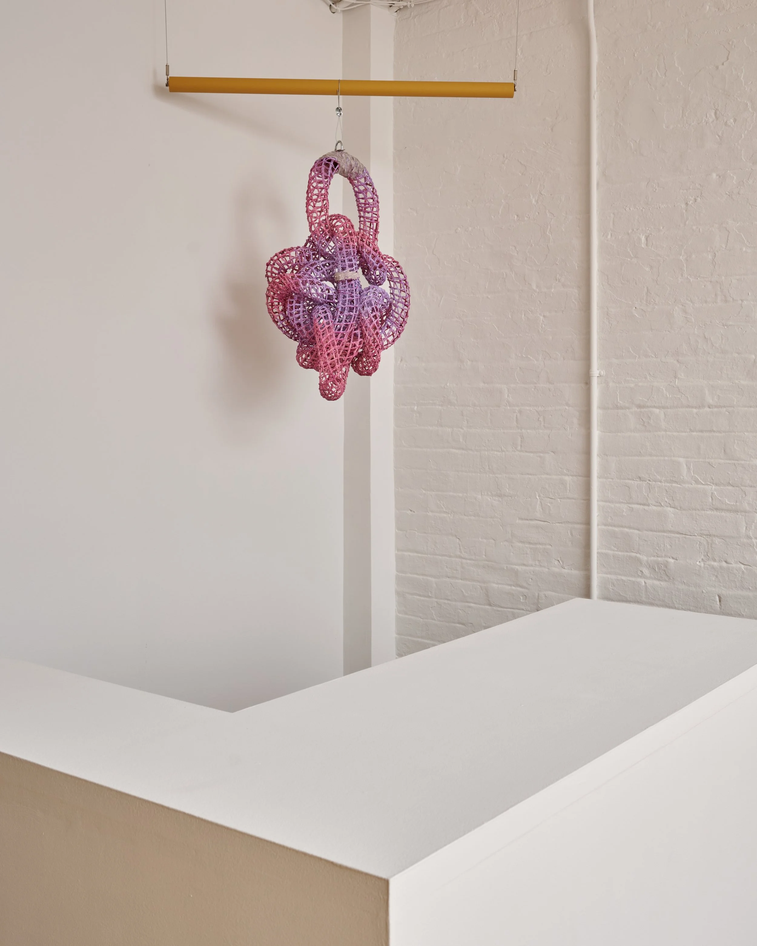 A pink and purple woven bag hanging from a yellow horizontal bar against a white wall with a brick texture.