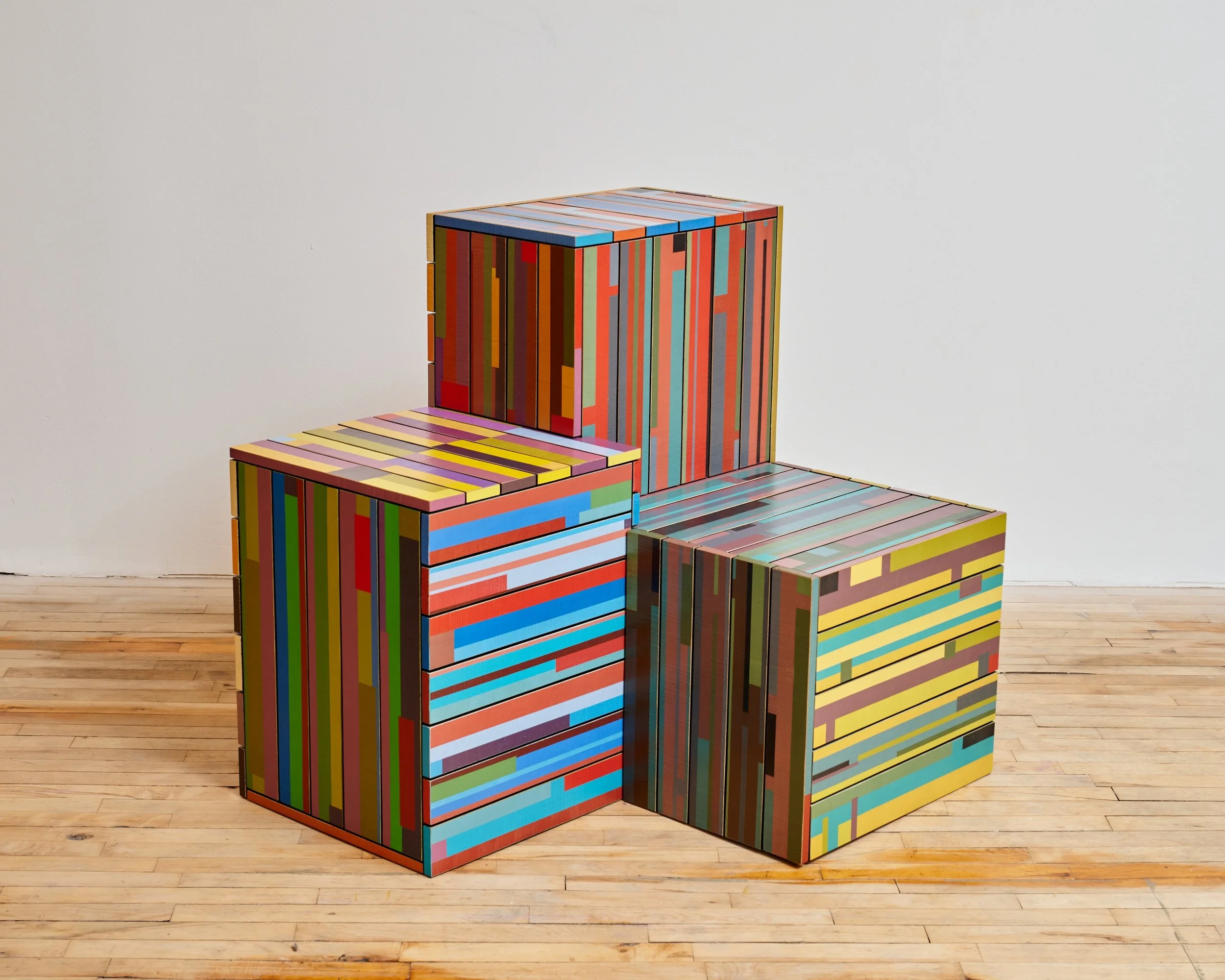 Three colorful, striped art boxes stacked in a corner of a room with wooden flooring and white walls.