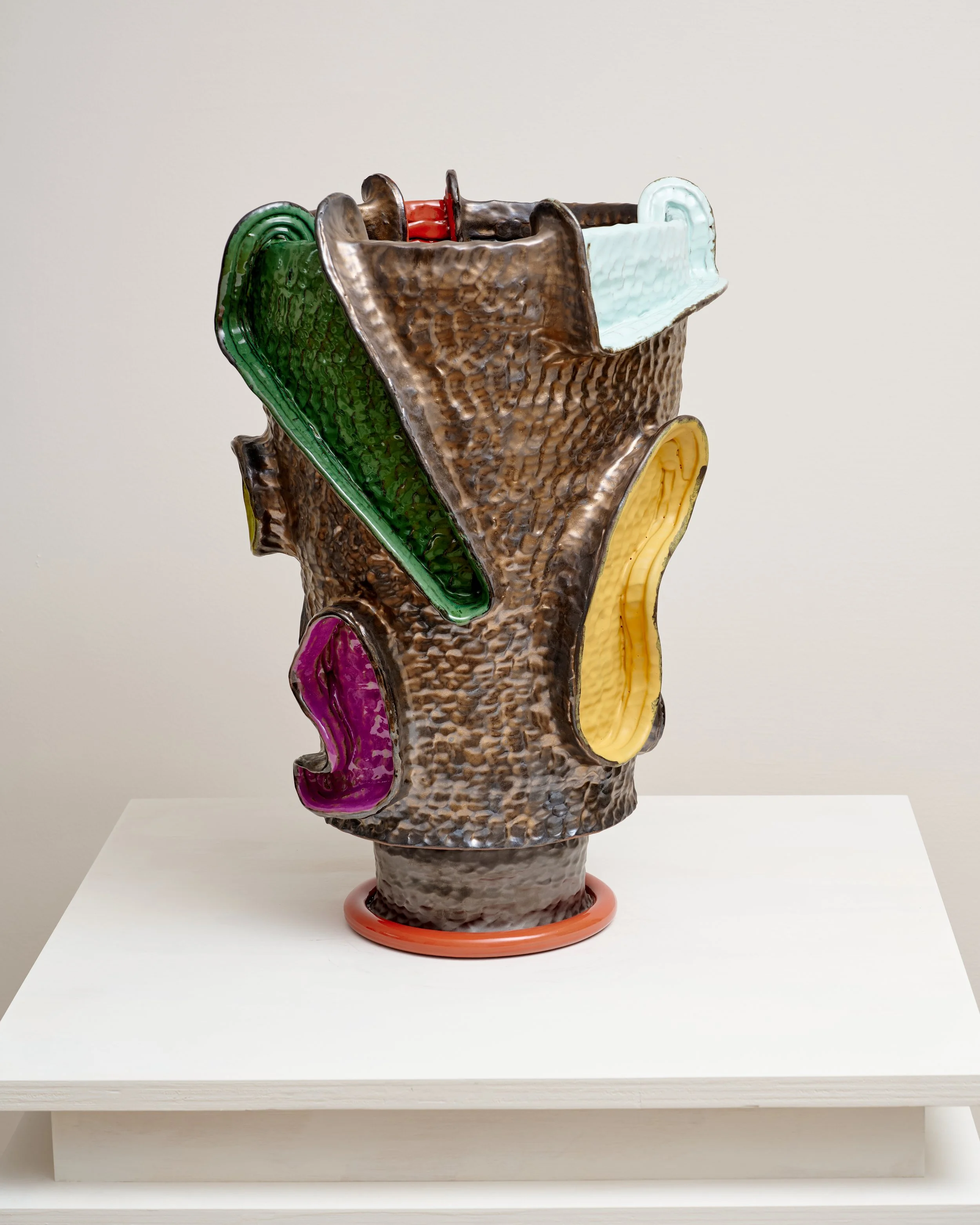 A textured ceramic sculpture resembling a tree trunk with colorful, irregularly shaped elements in green, purple, yellow, and light blue attached to its surface, placed on a white pedestal.