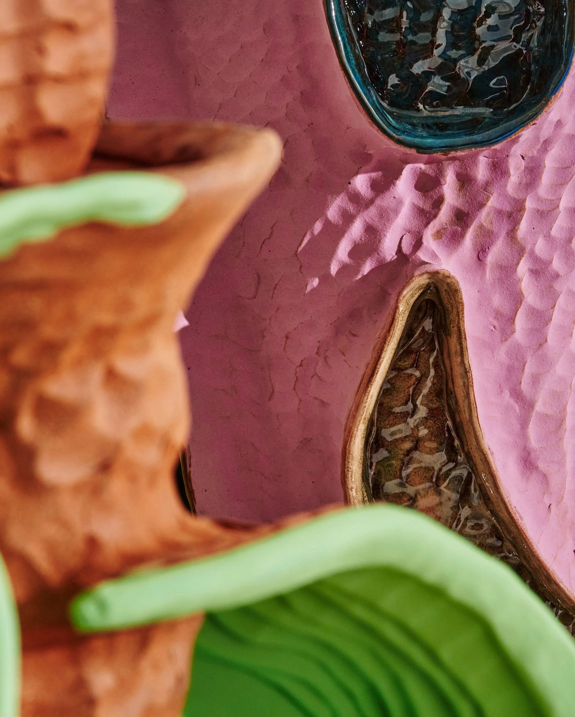 Close-up of a person wearing a light green top, with only part of their arm and shirt visible, near a textured pink surface, with a glass bowl filled with dark pebbles and a wooden sculpture in the background. Sean Gerstley.