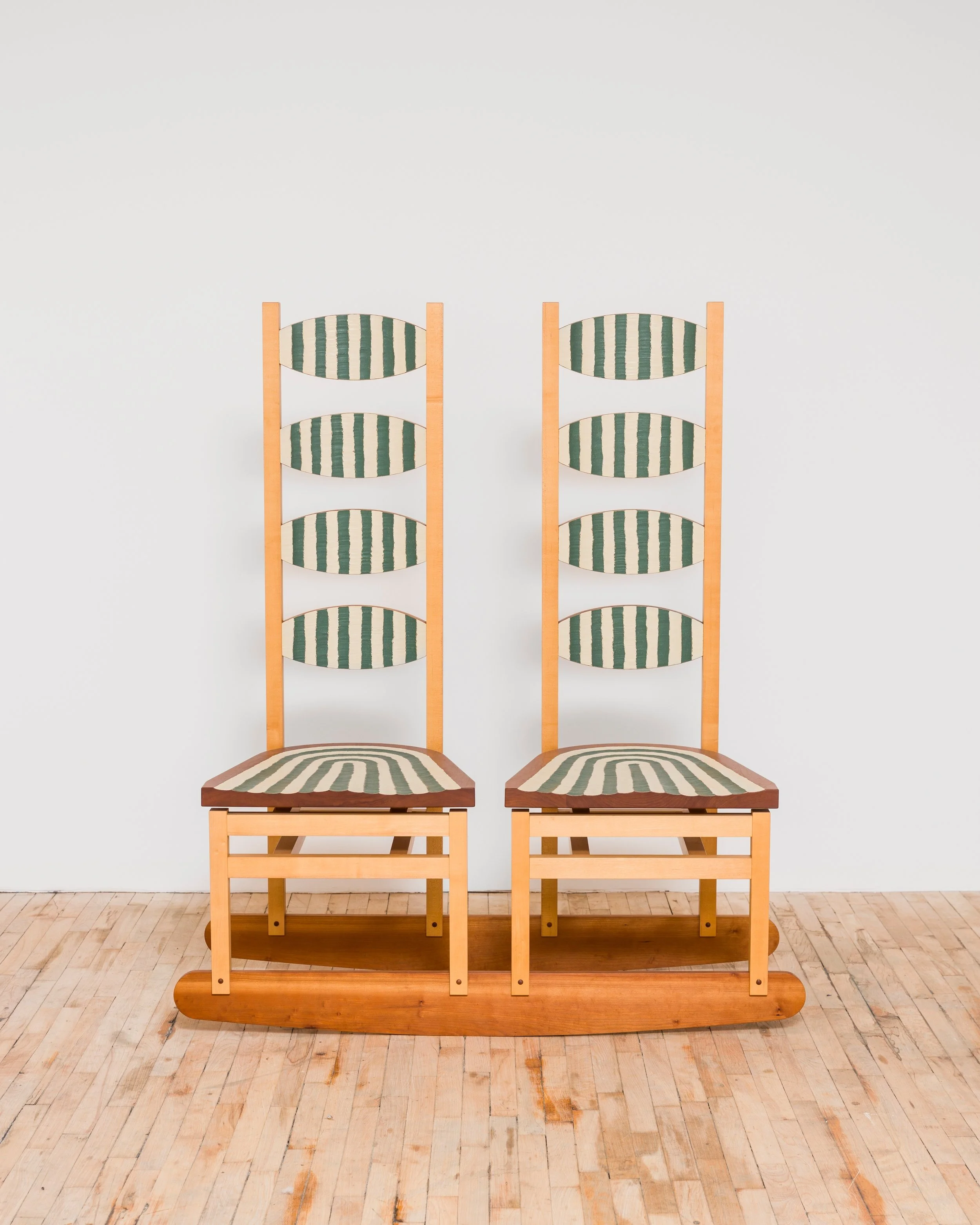 Two wooden chairs with high, oval-shaped backrests and striped green and beige cushions, placed side by side against a plain white wall on a wooden floor by Tom Loeser.