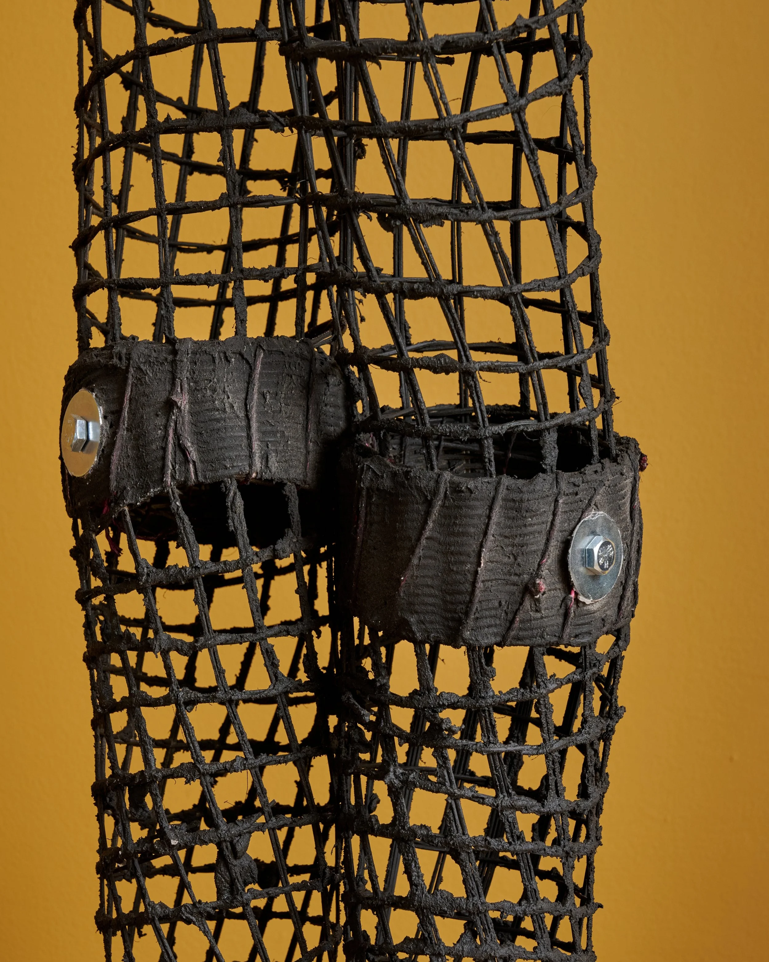 Close-up of a metal wire sculpture with two black straps attached on a yellow background.