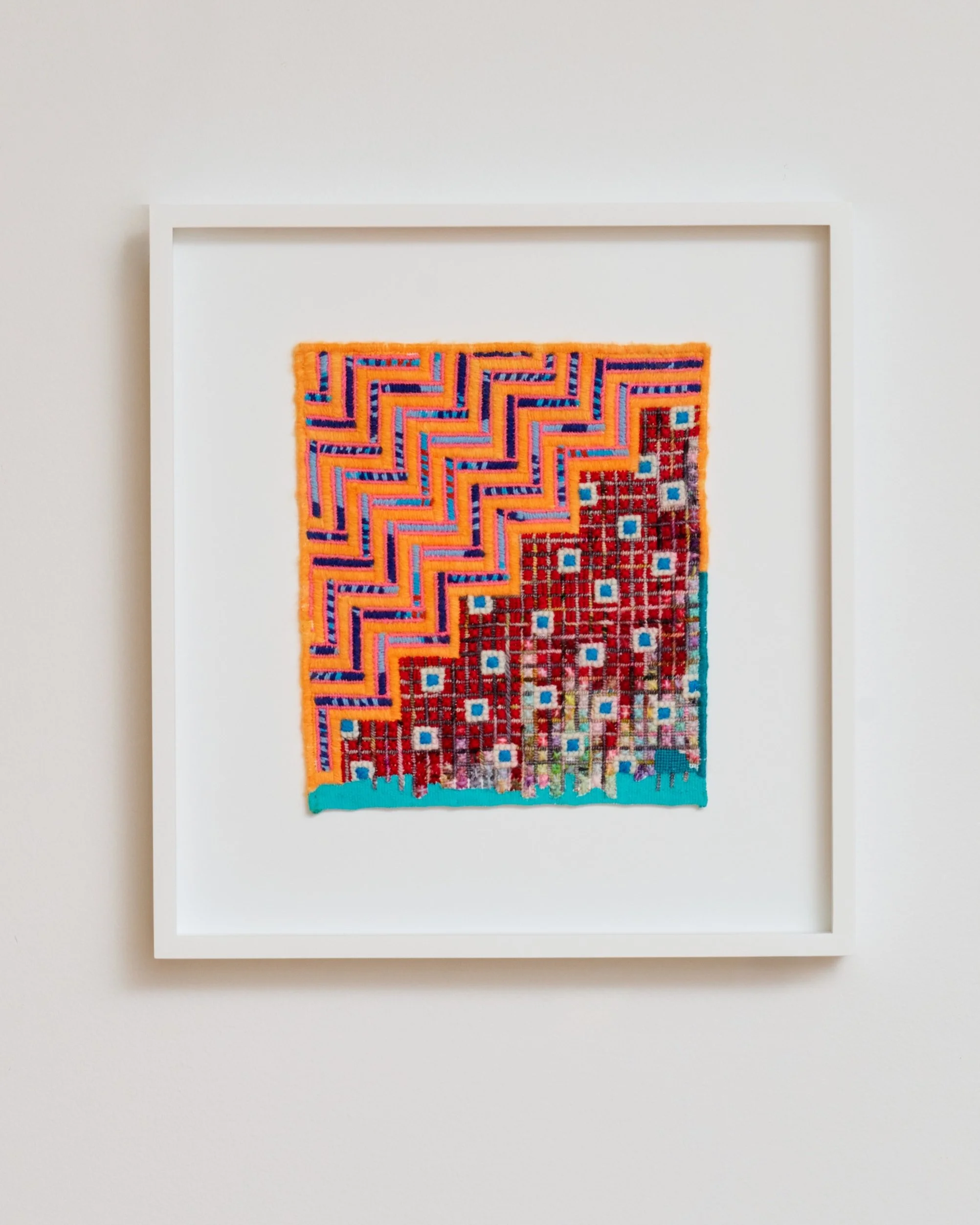 Colorful embroidered textile art featuring geometric patterns and cityscape silhouette, framed on a white wall.