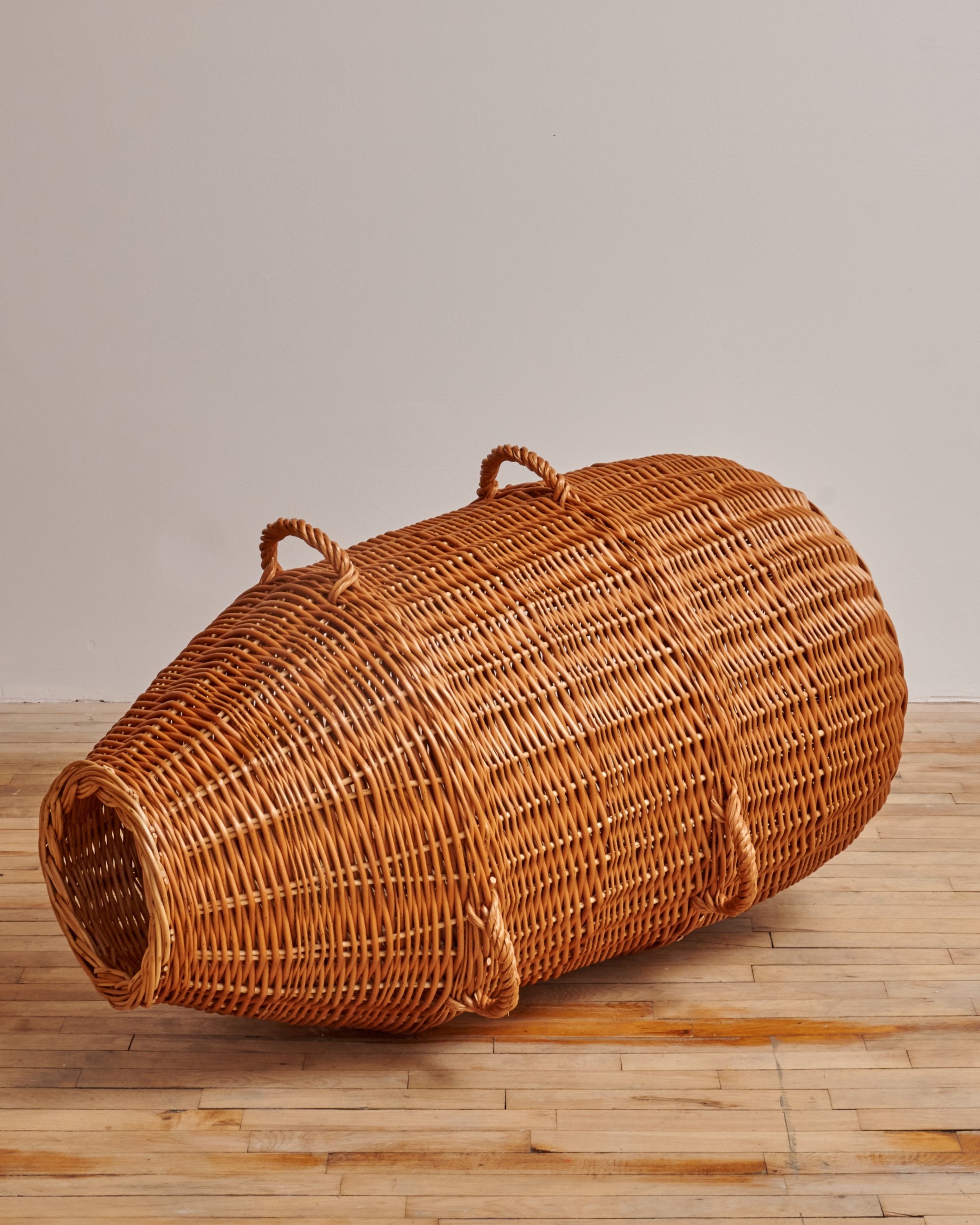 Wicker sculpture resembling a large bottle on a wooden floor against a plain wall.