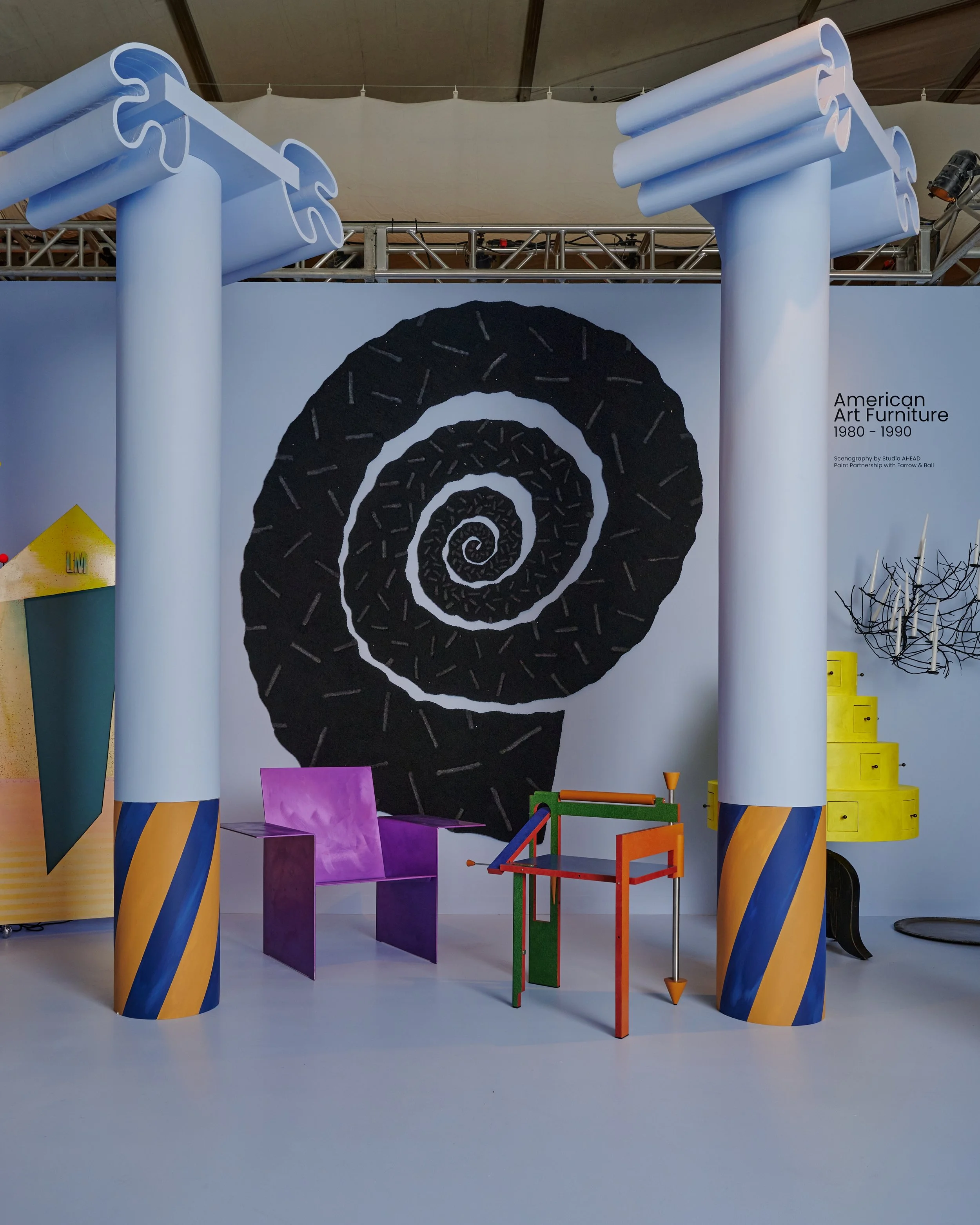An art exhibit showcasing modern furniture designs inspired by American art from 1980 to 1990, with colorful, abstract furniture pieces and a large black spiral artwork in the background.
