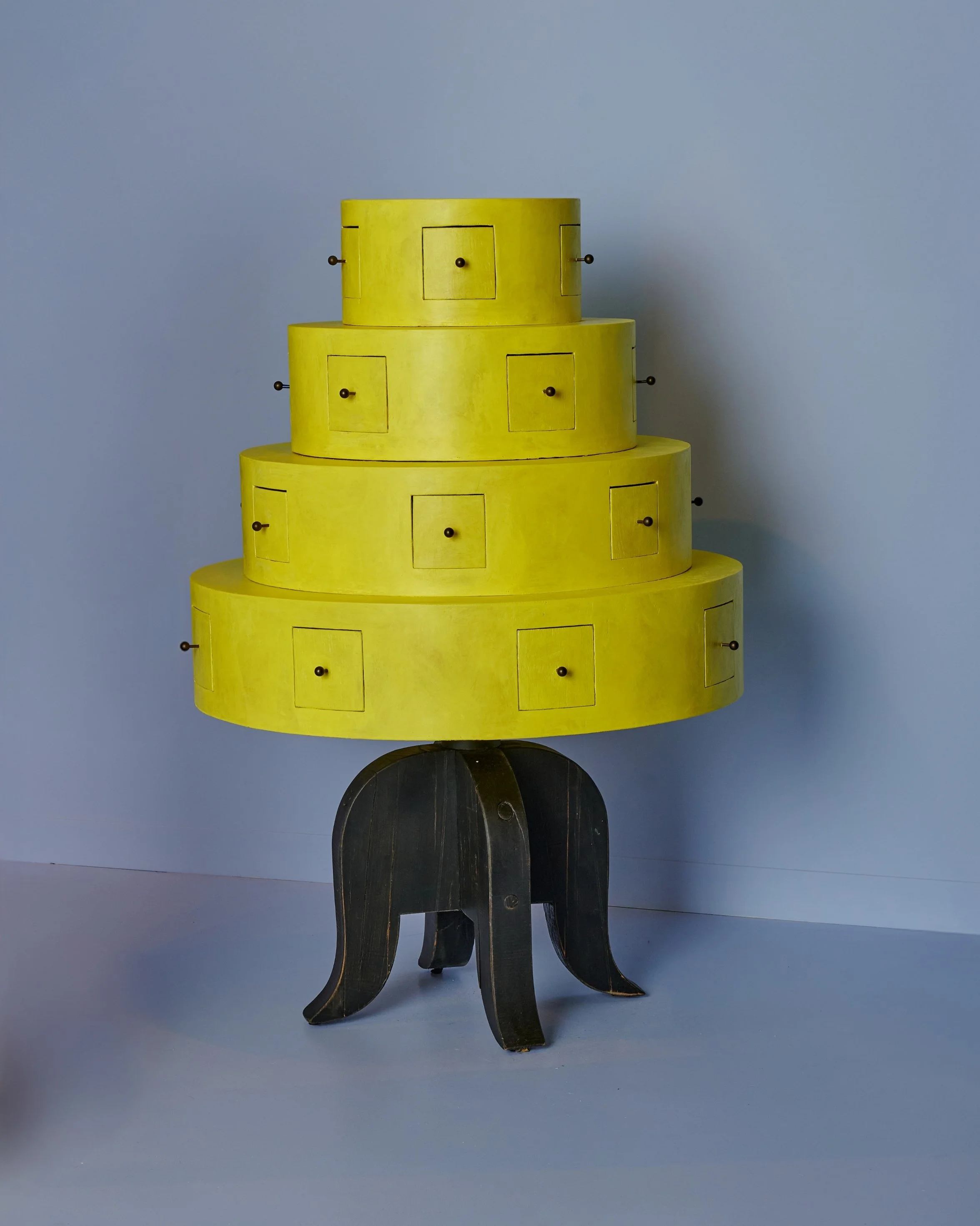 A yellow, tiered dresser with small drawers on each tier, mounted on a black, curved table base, set against a light blue background.