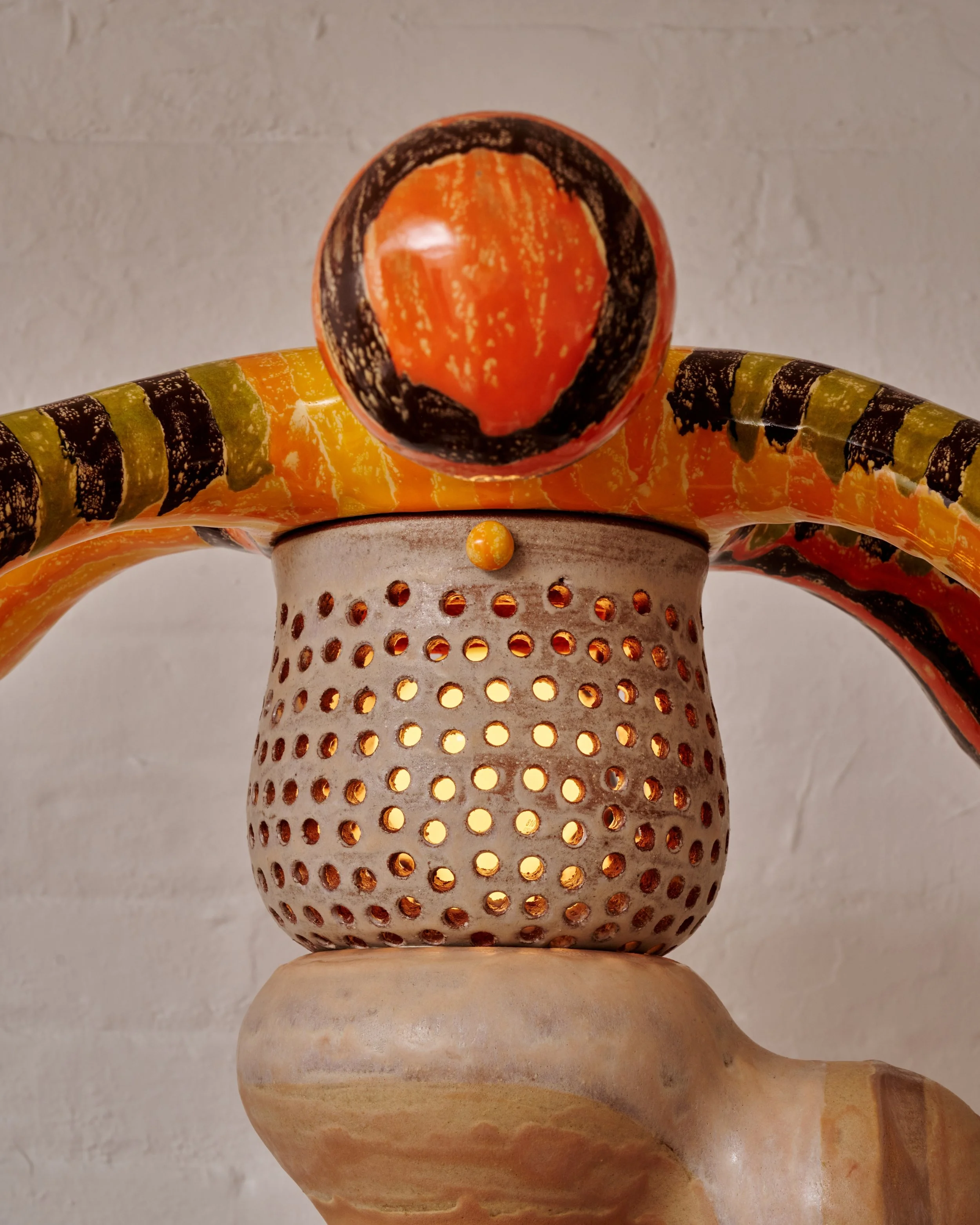 Close-up of a decorative metal object with a perforated pattern, featuring a round top with orange, black, and yellow colors, resembling a pumpkin or gourd, mounted on a carved wooden base. Joël Brodovsky-Adams.