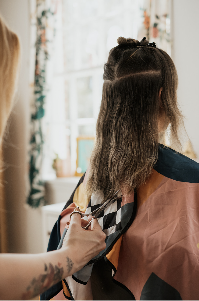 Woman getting her hair cut at Ceremony salon, seated with her shoulder uncovered by a colorful cape, while a hairstylist with tattoos cuts her dark brown hair with scissors.