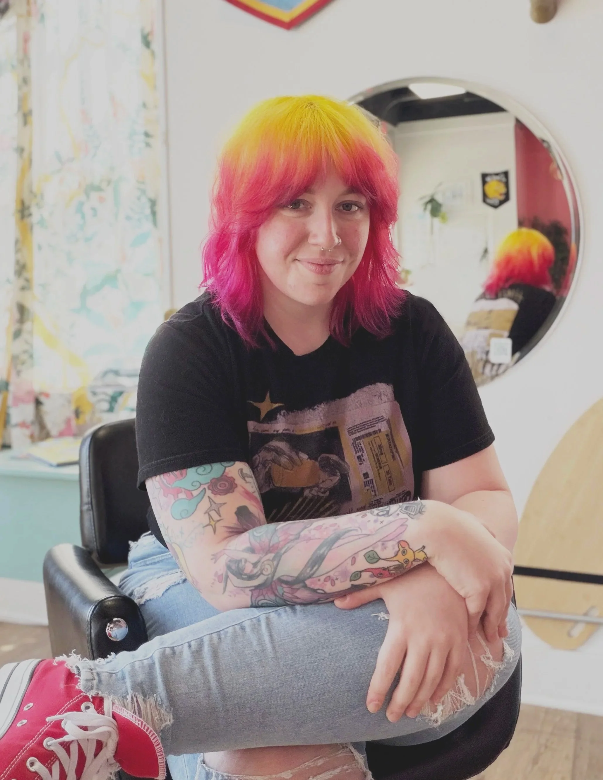 A young hair stylist with colorful pink and yellow hair, light skin, and earrings, wearing a black t-shirt and leopard print pants, standing indoors next to a white wall.