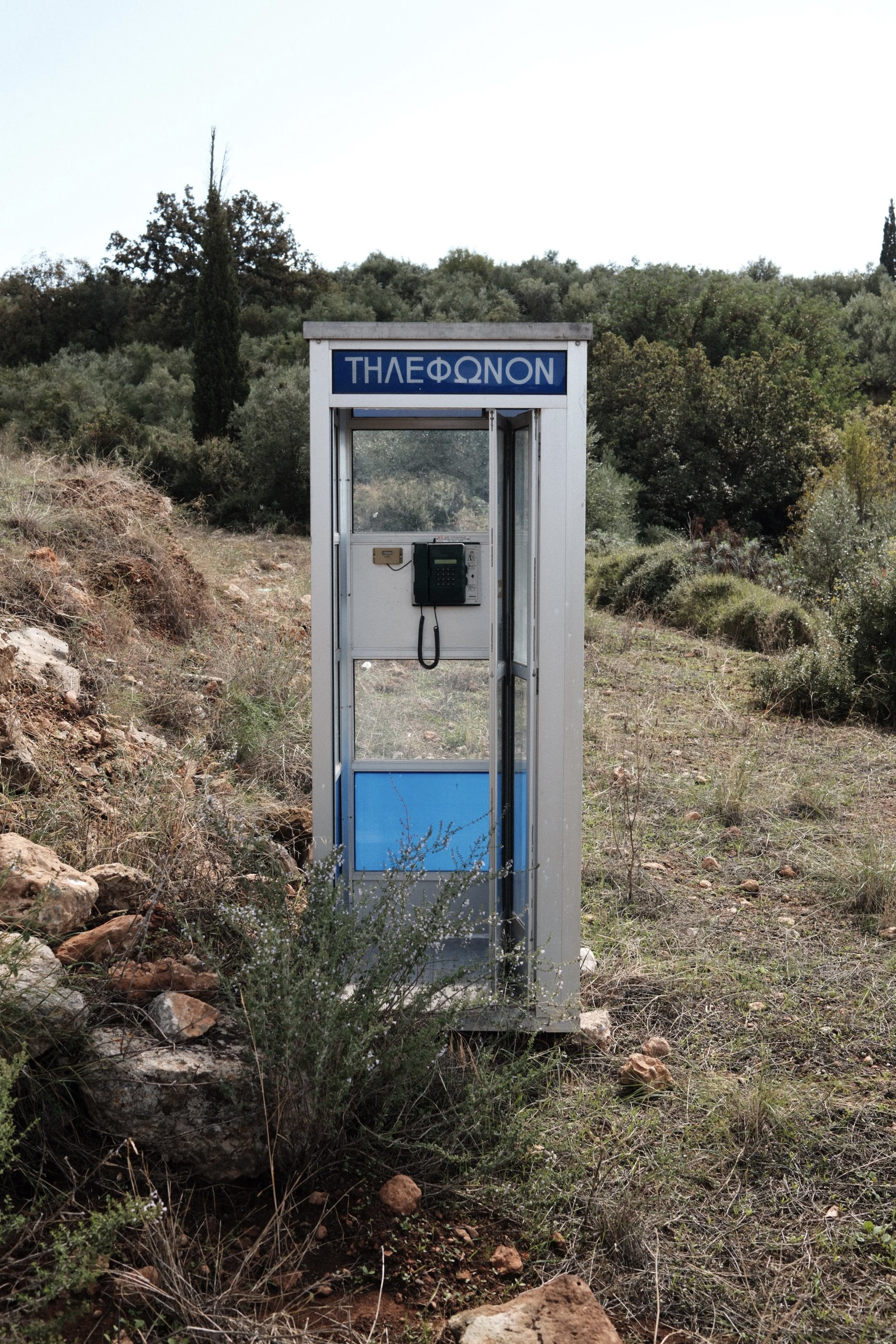 Empty outdoor telephone booth with Greek word 'ΤΗΛΕΦΩΝΟΝ' sign on top, situated in a natural landscape with grass, rocks, and trees.