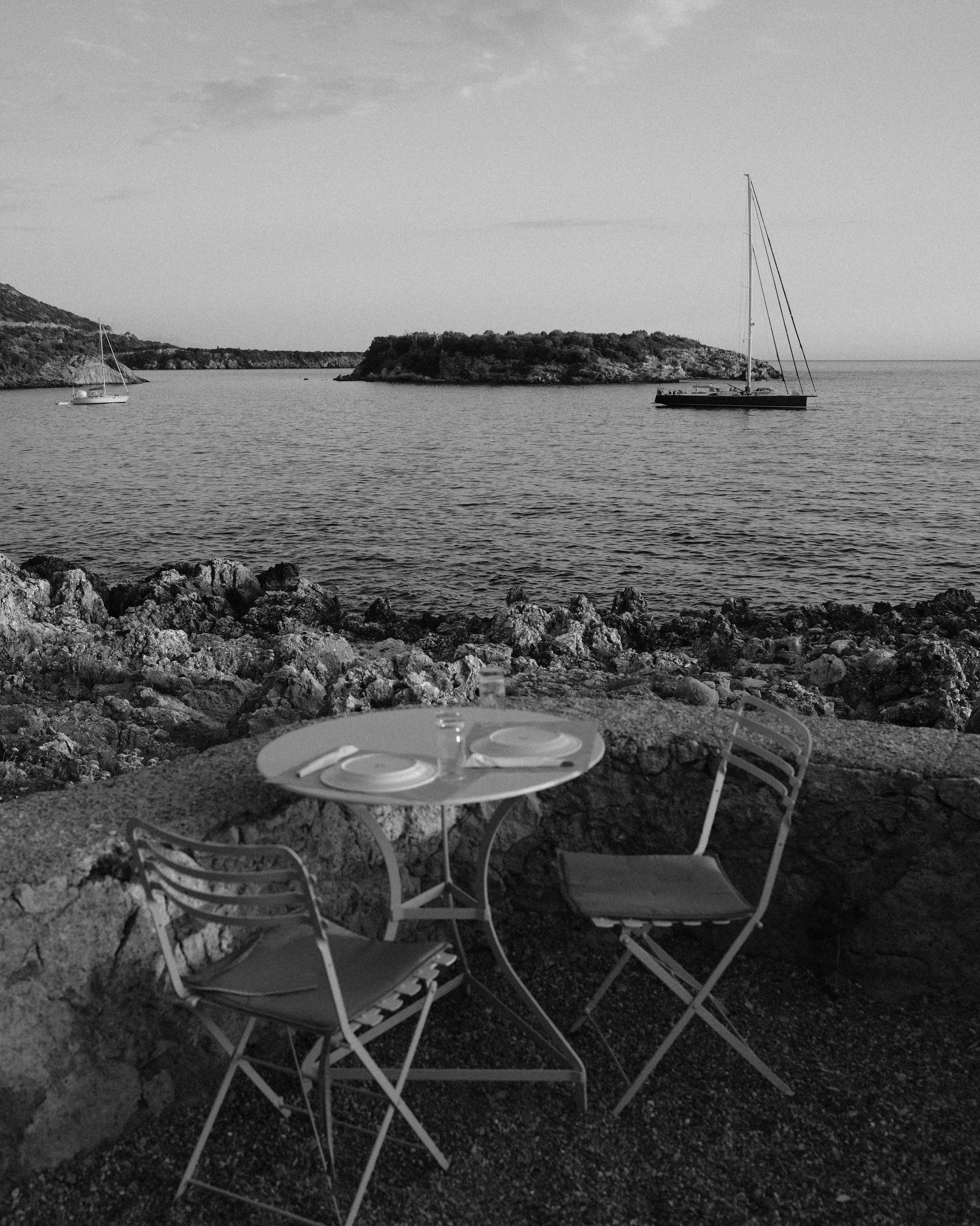 A black and white photo of a seaside view with two sailboats on the water, a rocky shoreline in the foreground with a small round table and two chairs, and islands in the distance under a cloudy sky.