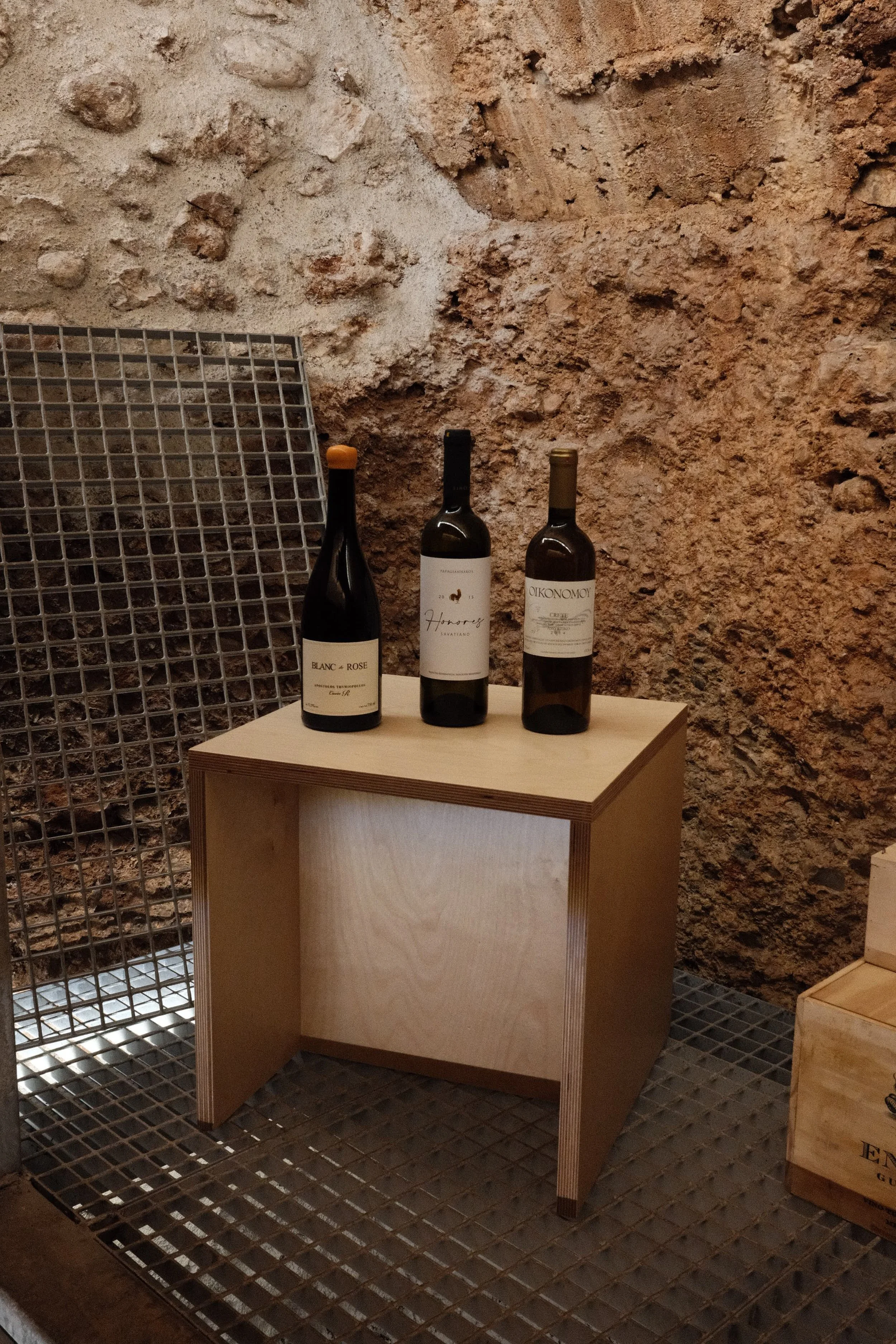 Three bottles of wine placed on a small wooden table against a rocky textured wall in a cellar or wine storage room.