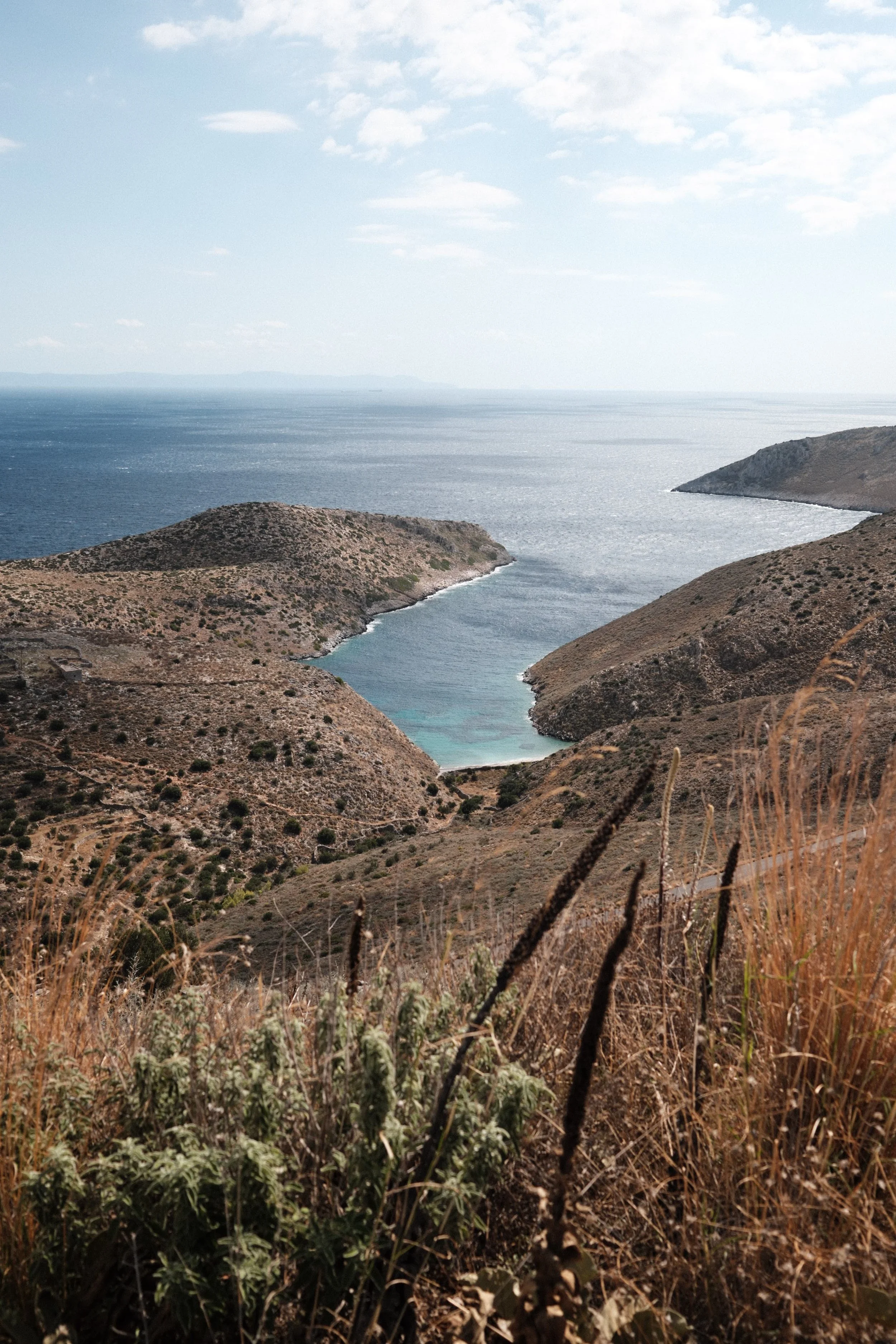 A view of a bay with a narrow inlet between hilly, dry landscapes leading to a calm ocean under a partly cloudy sky.