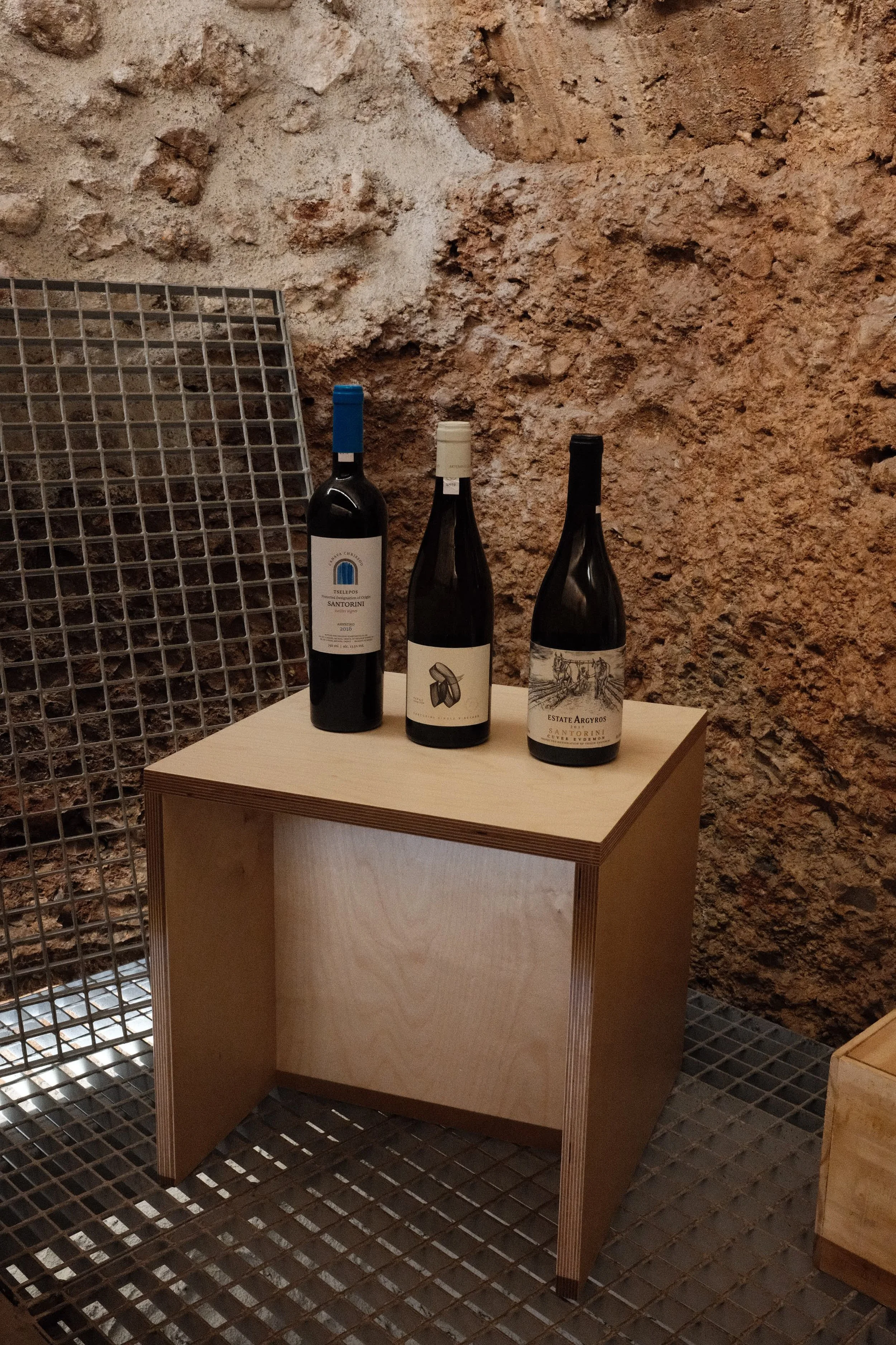 Three bottles of wine placed on a light wood display stand against a rough brick wall in a dimly lit environment.