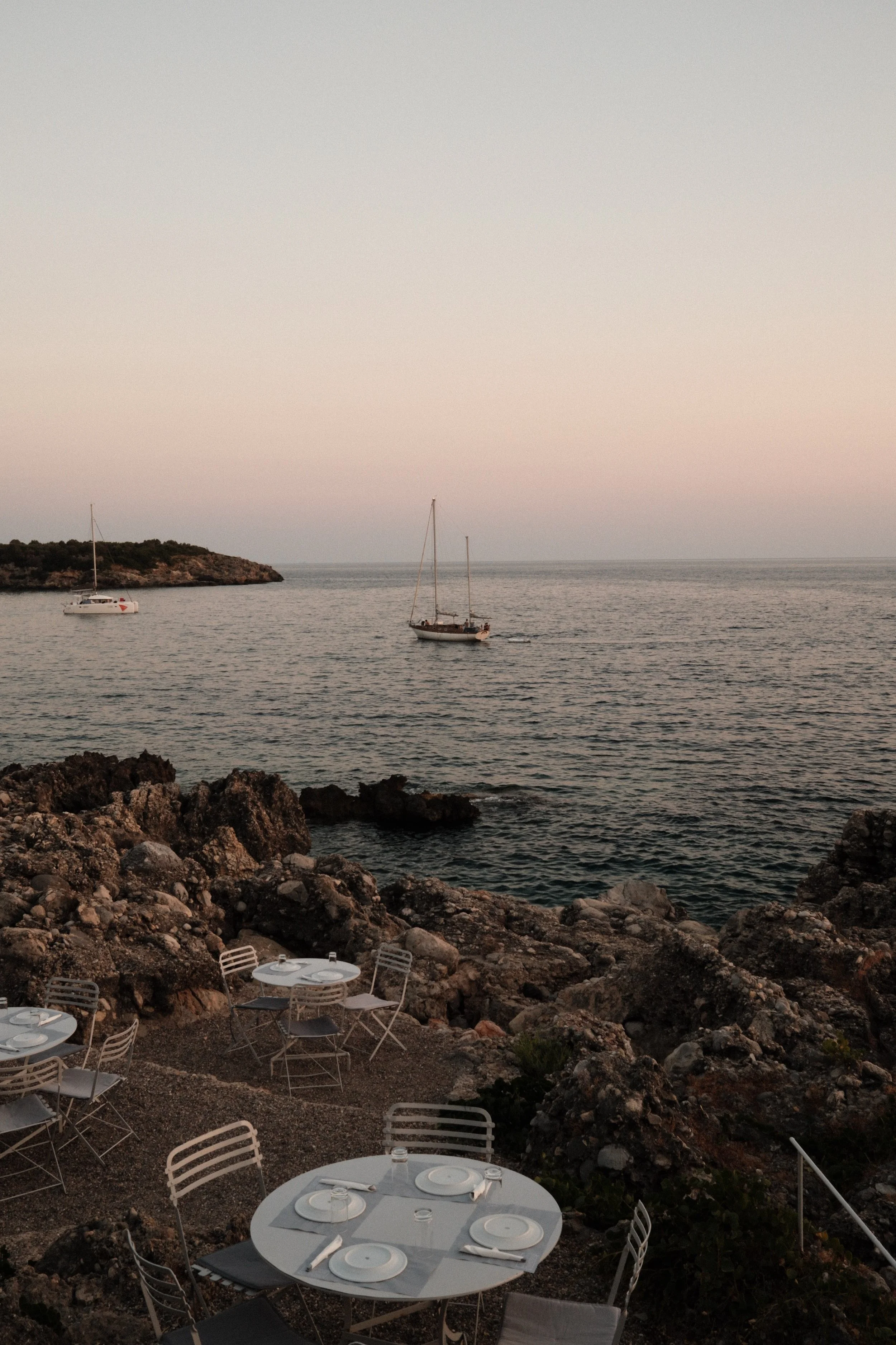 Outdoor dining area with tables and chairs set up on rocky terrain near the water's edge, overlooking the sea with two sailboats sailing nearby during sunset.