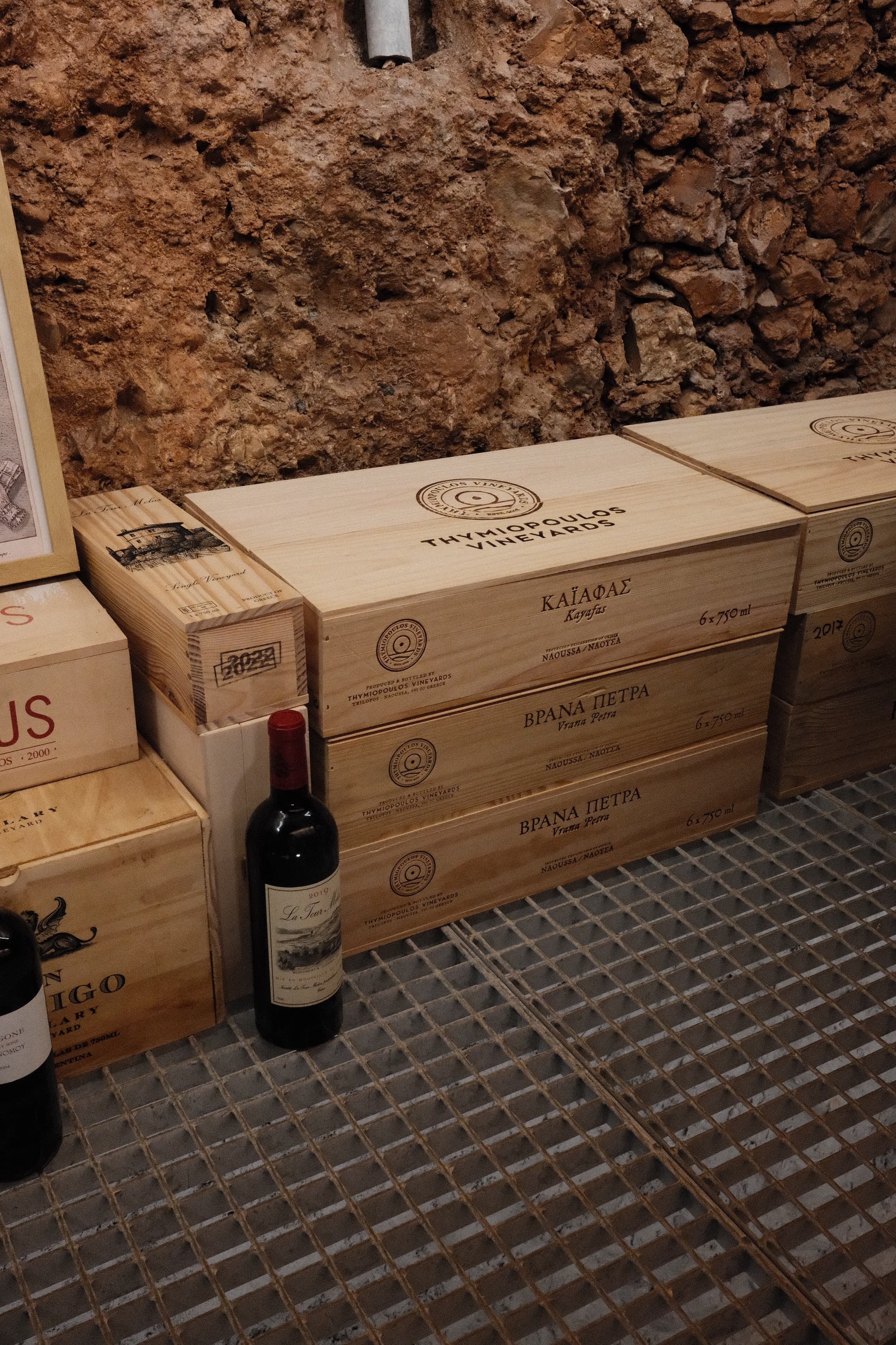 Wooden wine boxes and bottles displayed on a metal grid shelf against a textured brick wall.