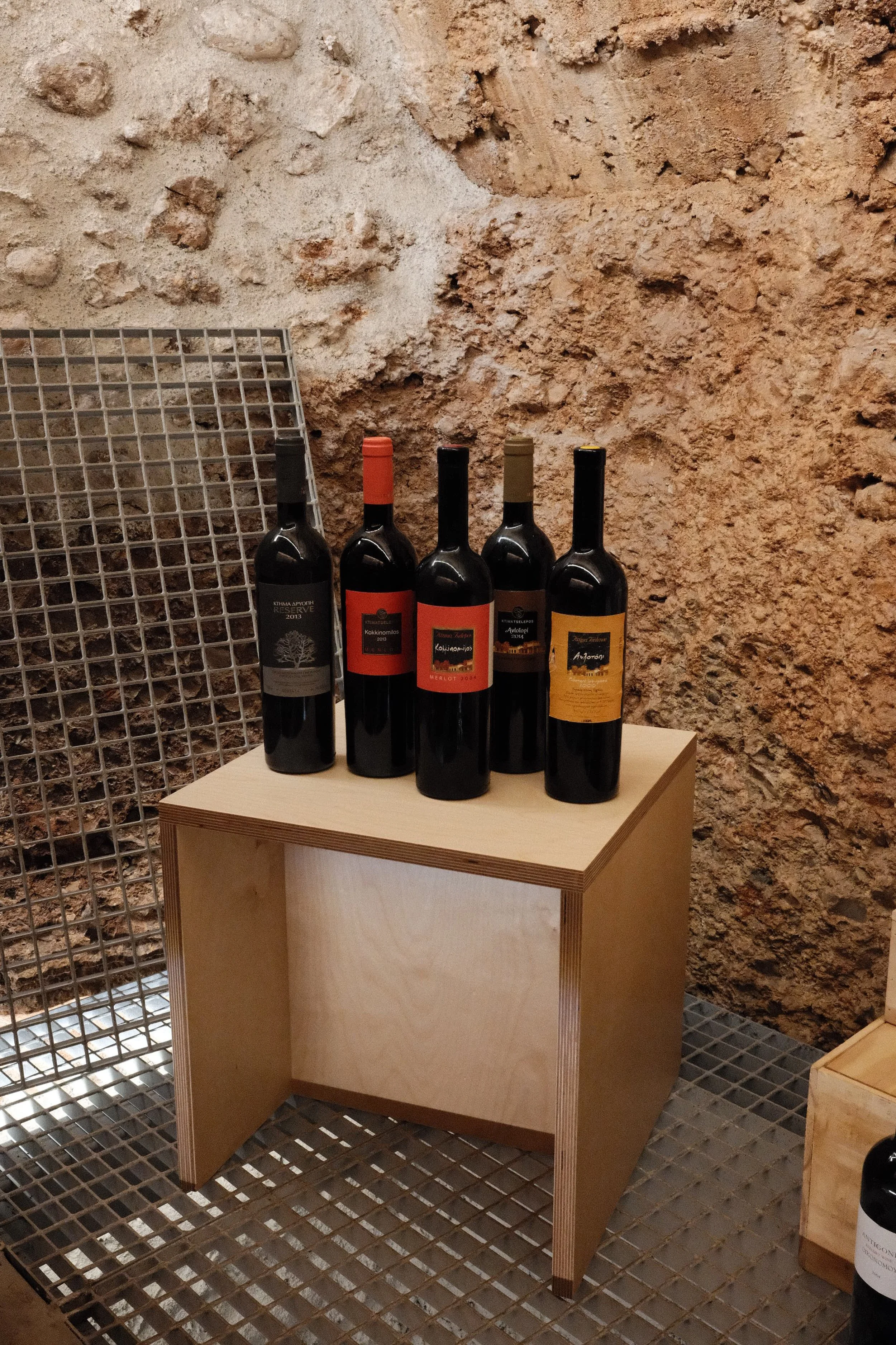Five bottles of wine are placed on a wooden table in front of a rustic stone wall, with a metal grid structure to the left.