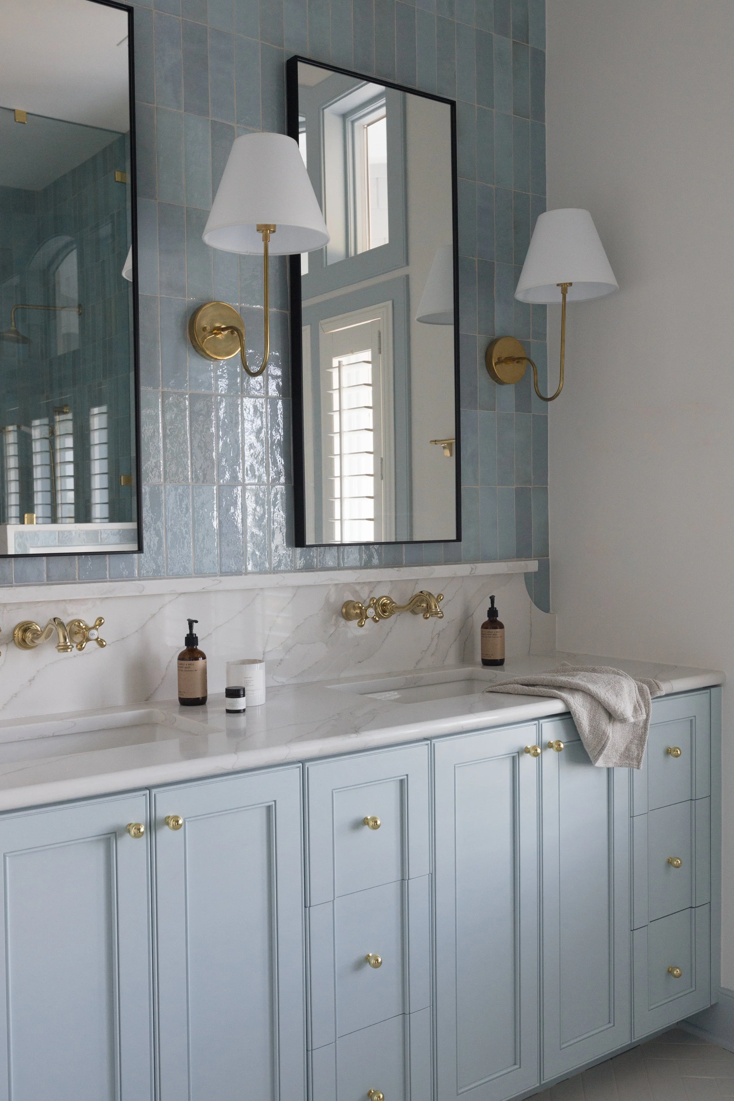 Bathroom vanity with a light blue cabinet, white marble countertop, two gold faucets, and a large mirror. Wall-mounted gold and white sconces flanking the mirror. Items on the counter include soap dispensers and small containers. A towel drapes over the right side of the counter.