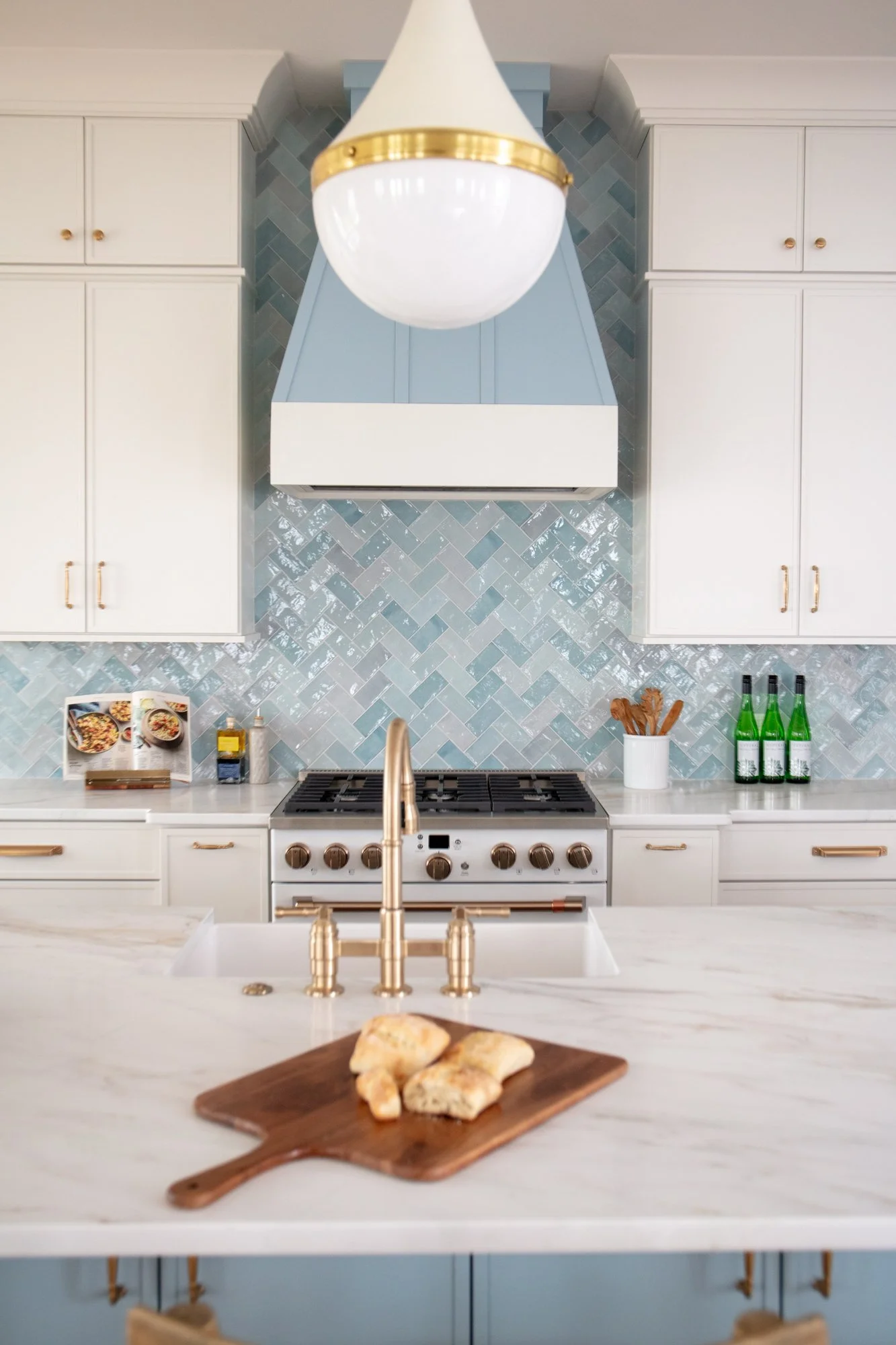 A modern kitchen with white cabinets, a blue and white range hood, a marble countertop, a gold faucet, and a decorative backsplash with a herringbone pattern. There are three green bottles on the counter, a white container with wooden utensils, a men