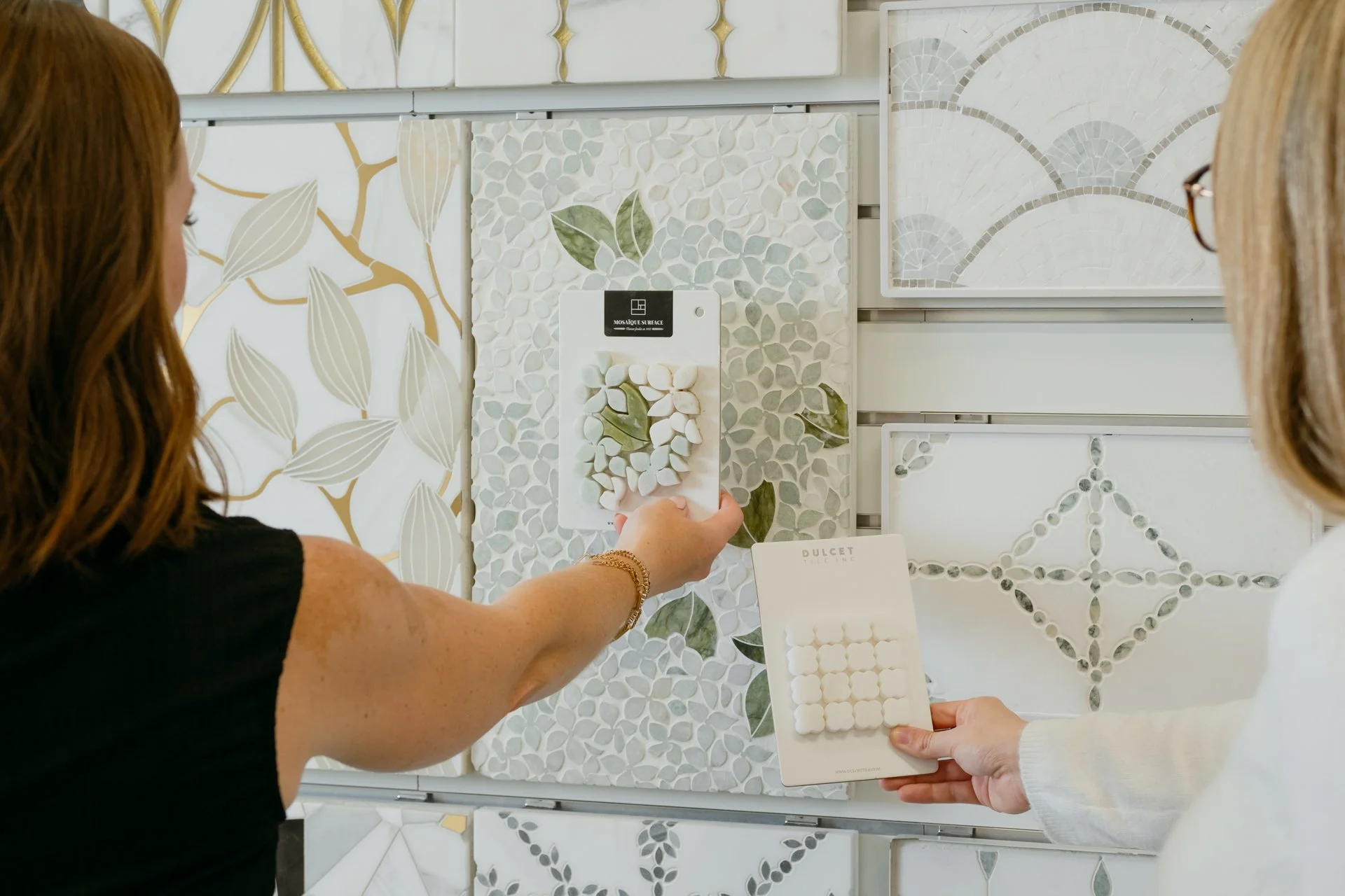 Two women are looking at a two samples of tile against a larger display of various colored and styled tiles at the Refine Tile and Design showroom in Wilmington, NC.