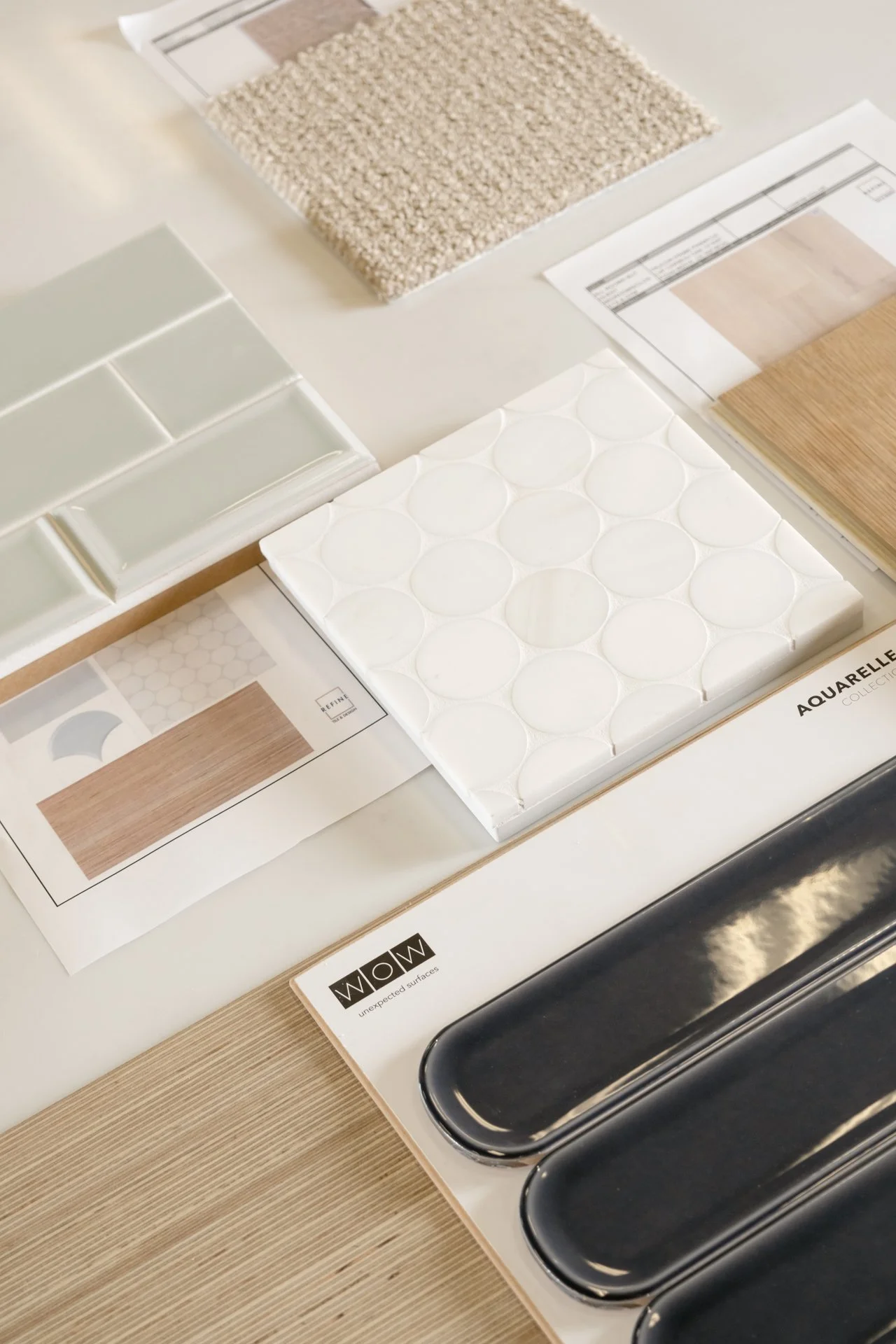 Various tile and flooring samples at the Refine Tile and Design showroom in Wilmington, NC including white ceramic tiles, beige textured tile, black geometric tile, and light colored wood and carpet samples.