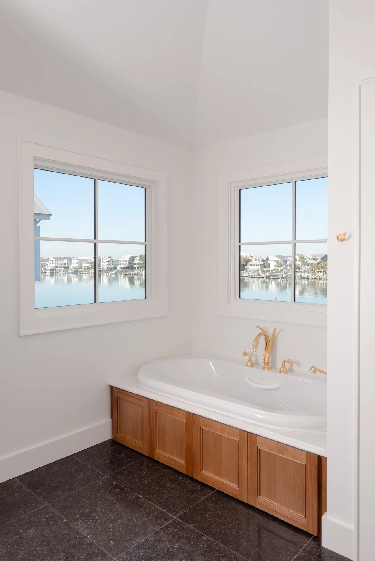 A master bathtub with a large format 12x24 dark charcoal tile surrounded by warm wood tones and beautiful coastal views with gold fixtures.