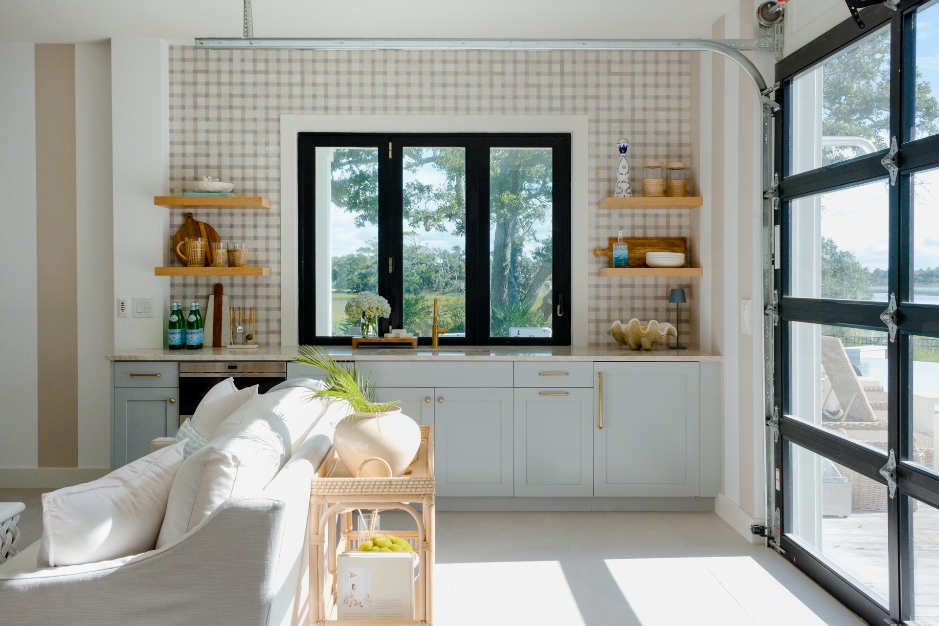 Bright kitchen with large window showcasing an outdoor view, white and gray cabinets, open wooden shelves with decorative items, and a sliding glass door to patio with outdoor furniture.