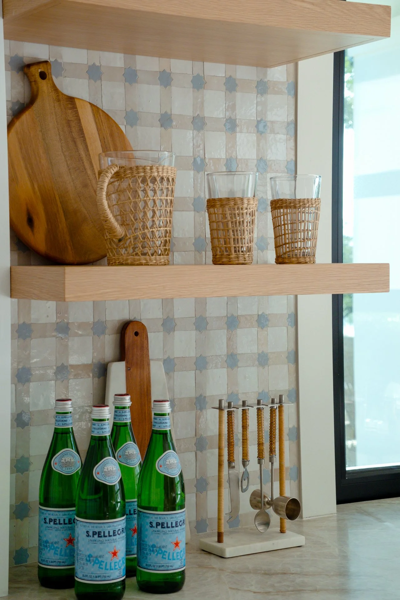 Kitchen shelf with a wooden cutting board, glassware, four glass bottles of sparkling water, and kitchen utensils.