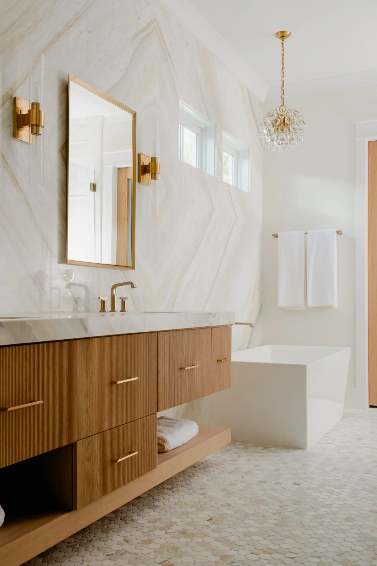 Bathroom with two sinks, gold fixtures, large mirrors, white countertops, white large slab walls, natural light, freestanding bathtub with mosaic tile floor.