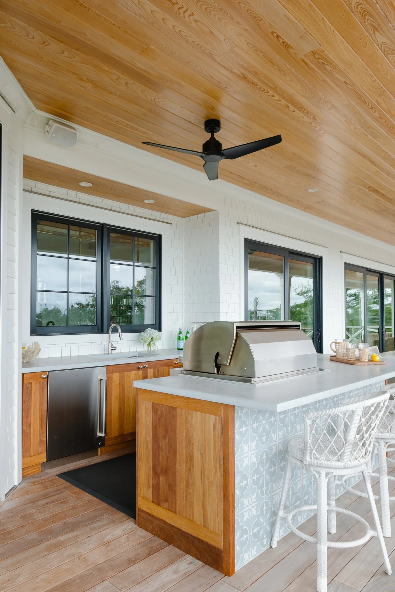 Outdoor kitchen with wooden cabinets, a stainless steel grill on a white counter, black framed windows, white wicker chairs, ceiling fan, and sliding glass doors.