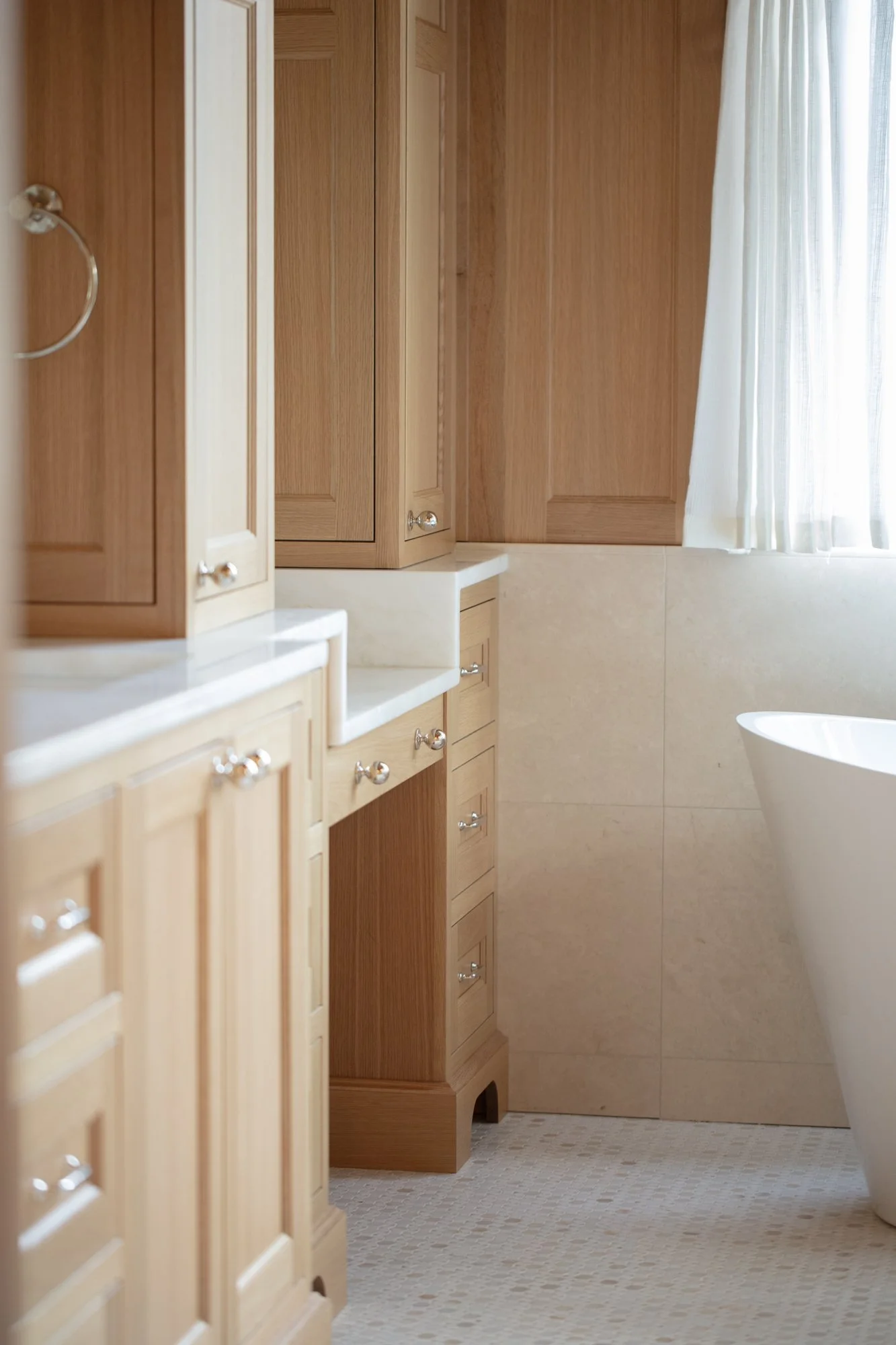 A bathroom with wooden cabinets, a marble countertop, beige tiled walls, a window with white curtains, and a white freestanding bathtub.