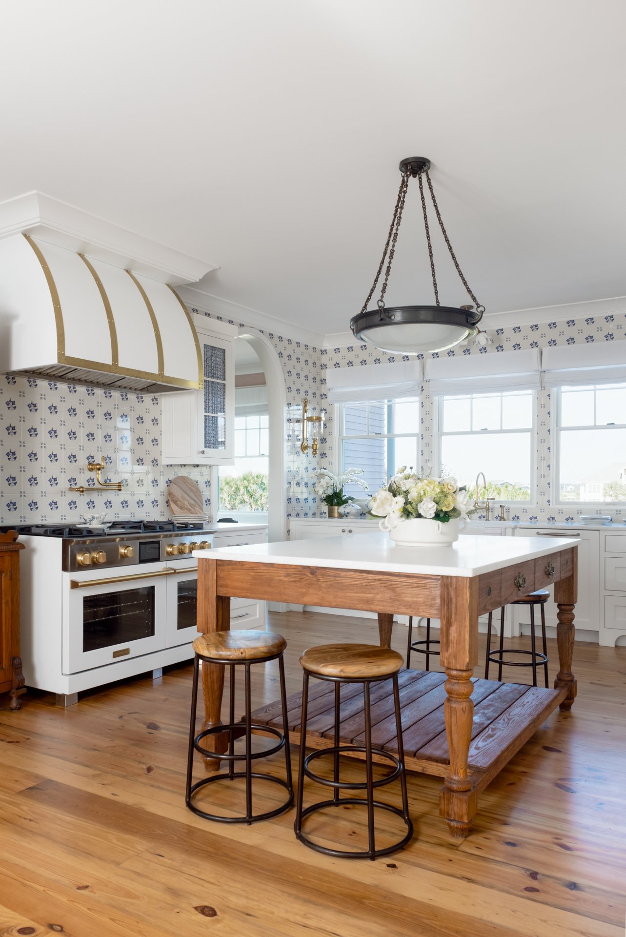 Bright kitchen with white cabinets, patterned blue and white wallpaper, a wooden kitchen island with a white countertop, three wooden and metal bar stools, large windows, and a chandelier.
