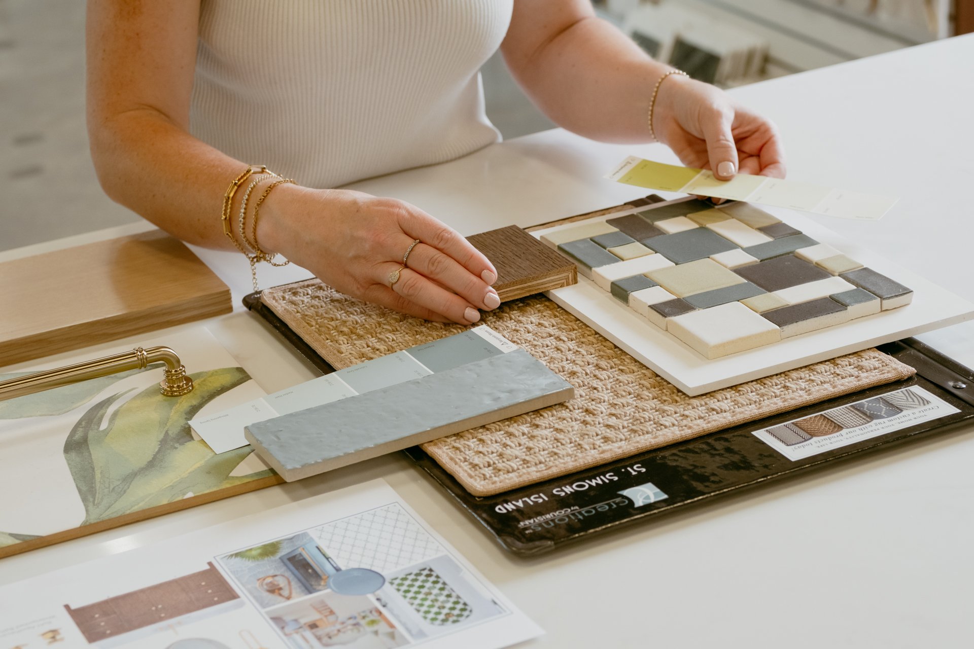 A flat lay of various color tiles, finishes, a carpet and hardwood sample with a women holding a paint sample at the Refine Tile and Design showroom in Wilmington, North Carolina.