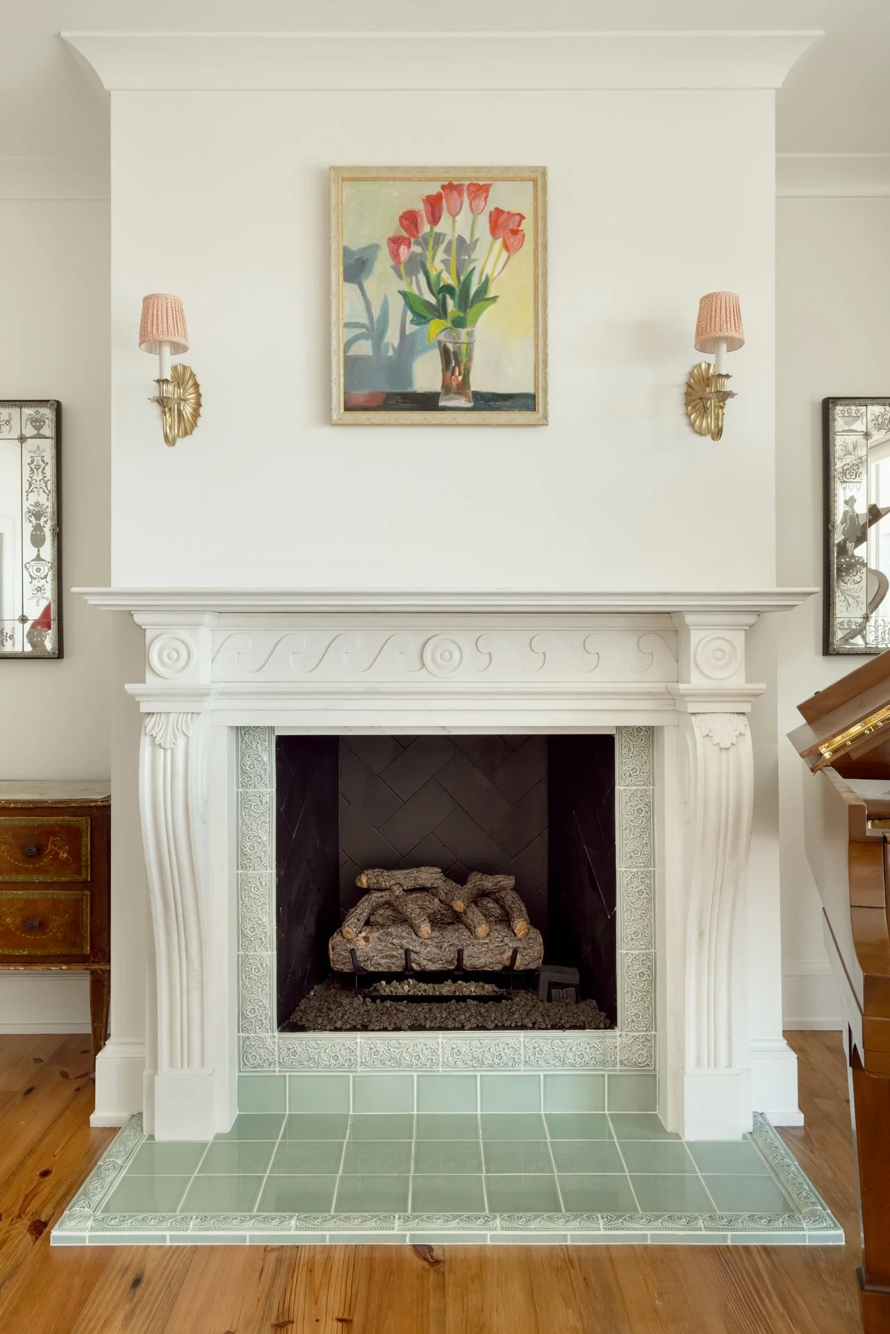 A white fireplace with detailed molding and black interior, topped with a framed painting of pink tulips in a glass vase. There are two pink wall sconces on either side of the painting, and part of a wooden cabinet and a piano are visible.