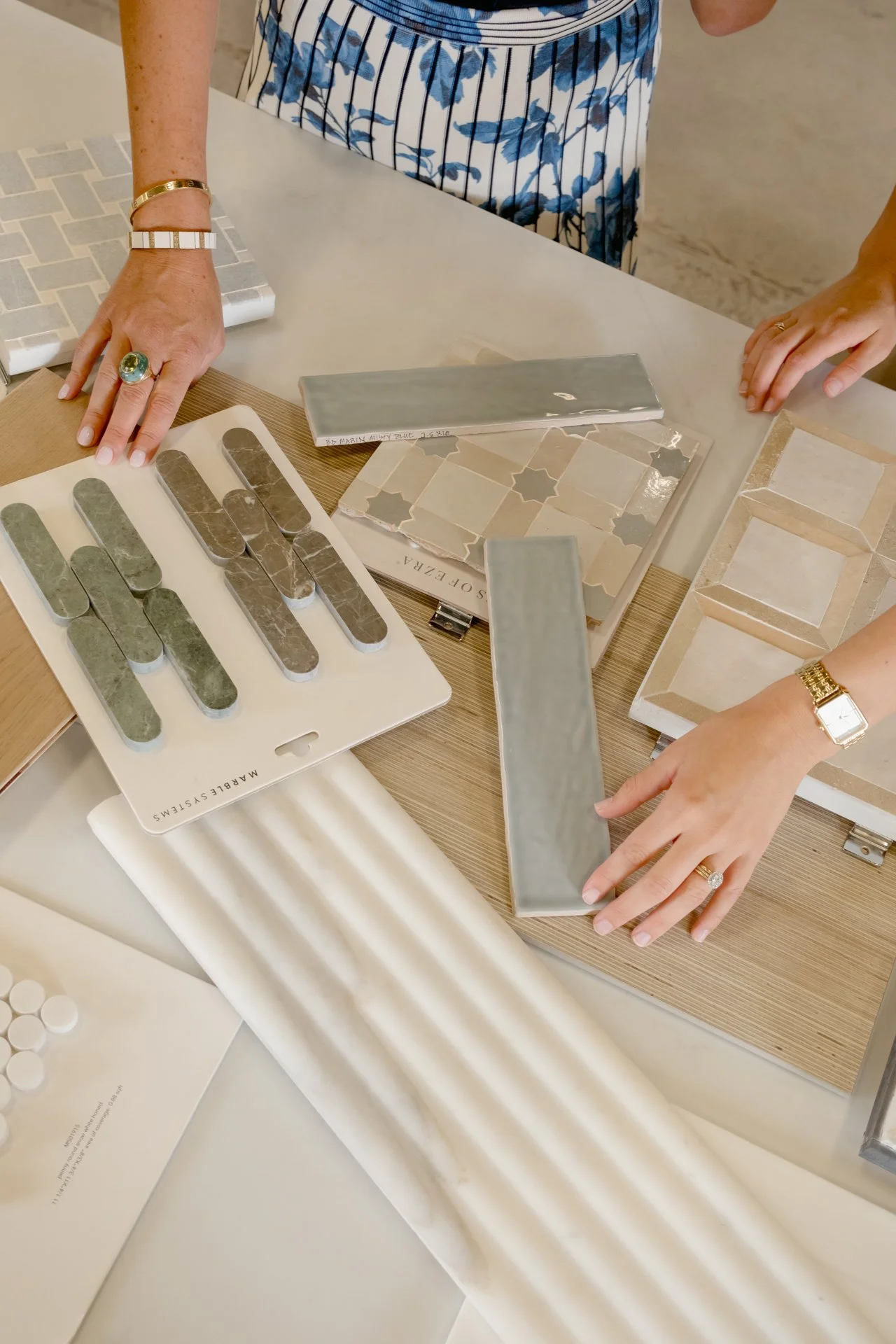 Design consultation at the Refine Tile and Design showroom in Wilmington, NC. Two people looking at various tile samples on a table, including small tiles in different colors and styles, and larger tiles in neutral tones.