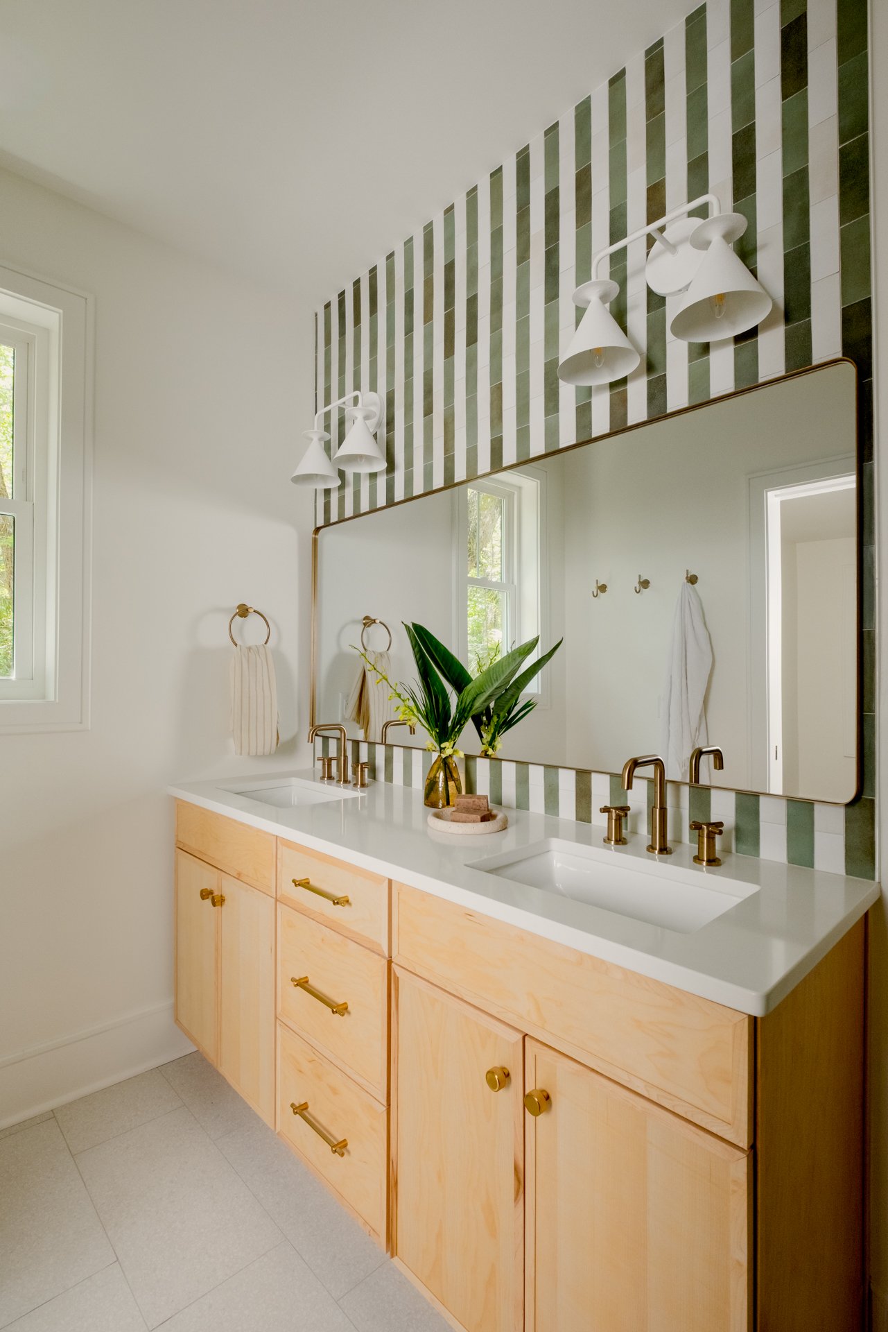 A bathroom with light wood cabinets, a white marble countertop, a full-size wall mirror, gold fixtures, white wall-mounted lamps, and a green and white stripe backsplash tile.