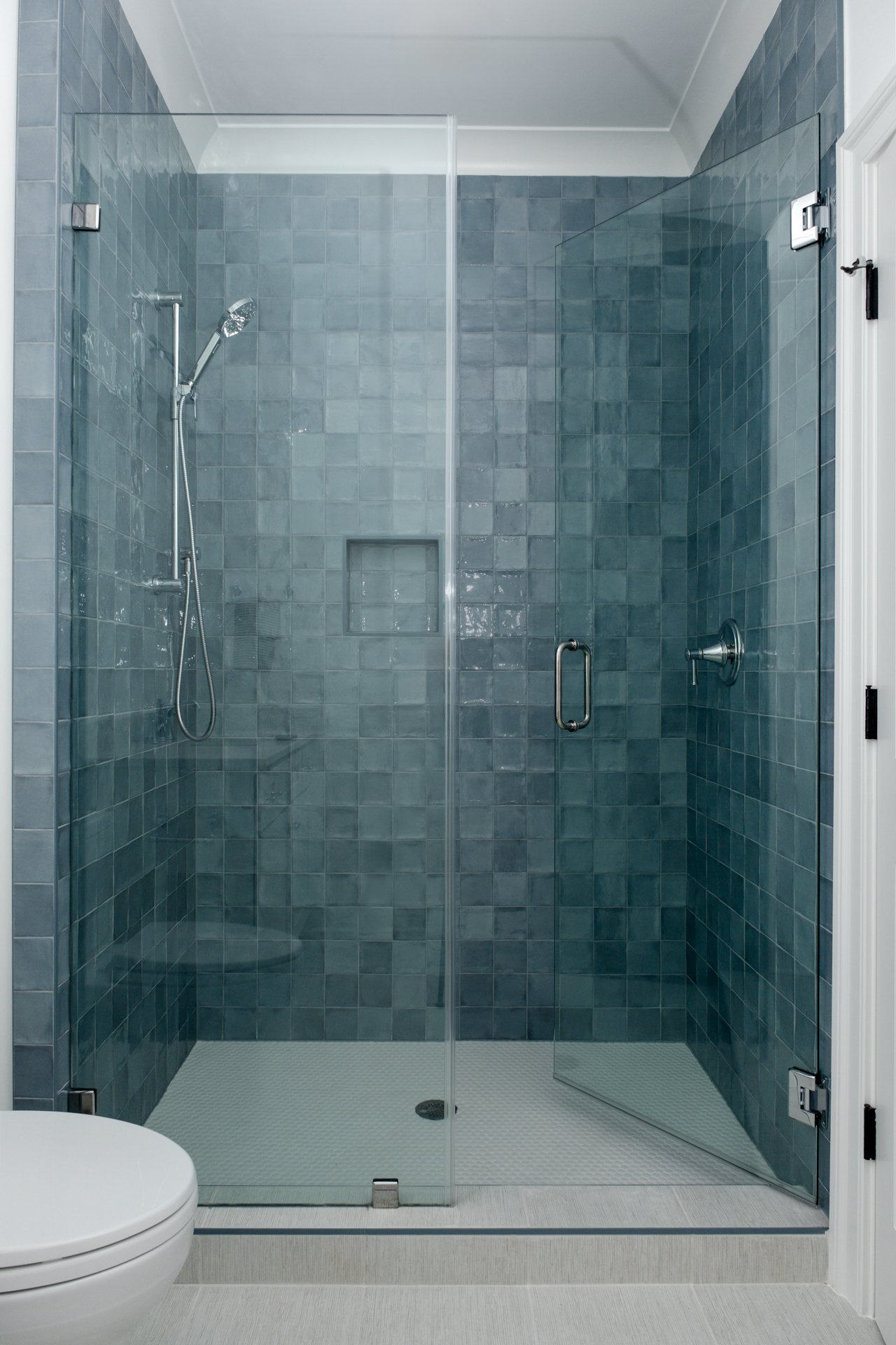 A glass shower enclosure with tiling inside, a handheld showerhead, and a built-in niche, with a white toilet partially visible in the foreground.