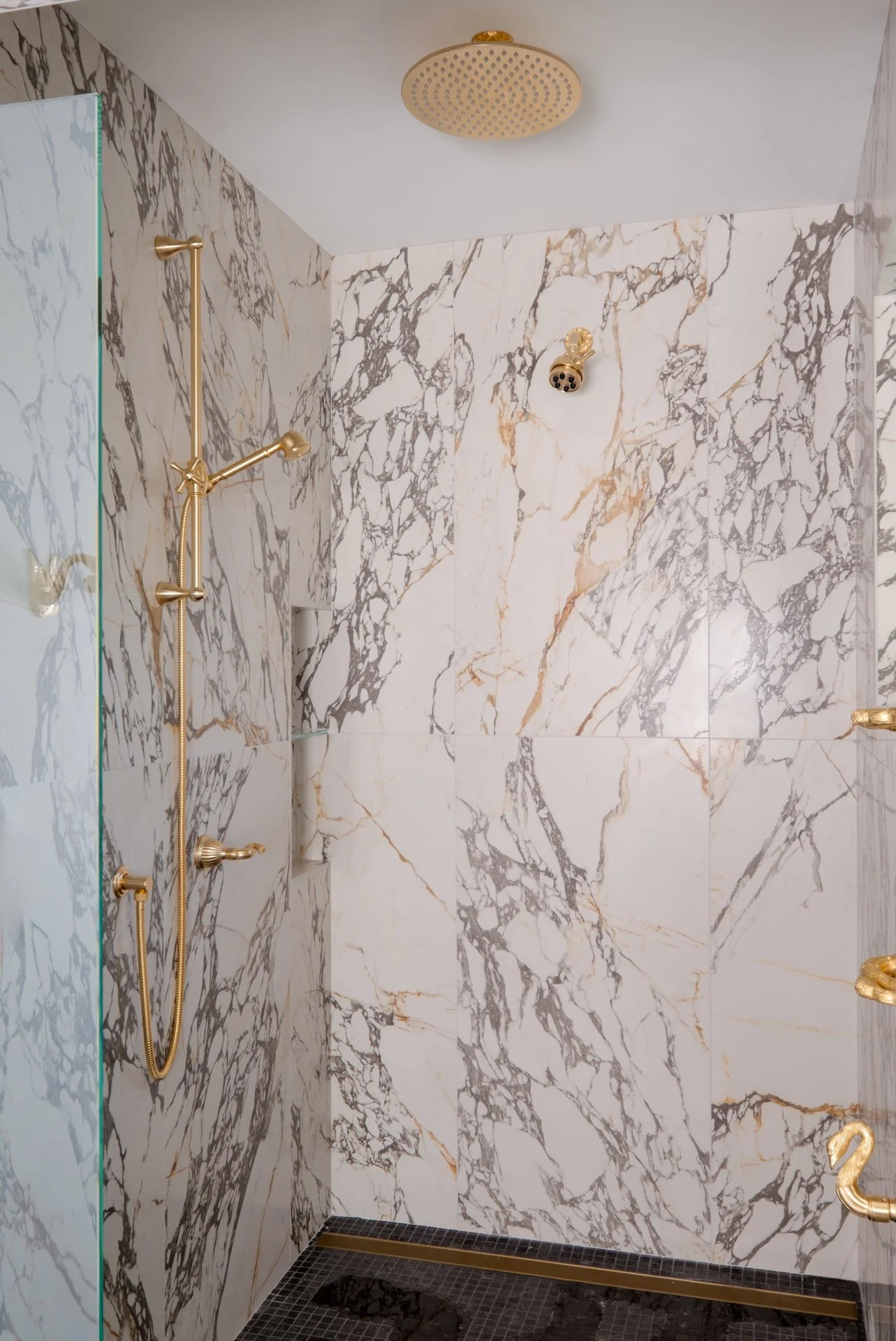 A large format master shower with Nemo Tile Leon Arabescato in 24x48 — Bold veining, timeless movement, and a scale that makes the entire space feel elevated and intentional.  Gold shower fixtures to pull out the gold in the veining of the tile.