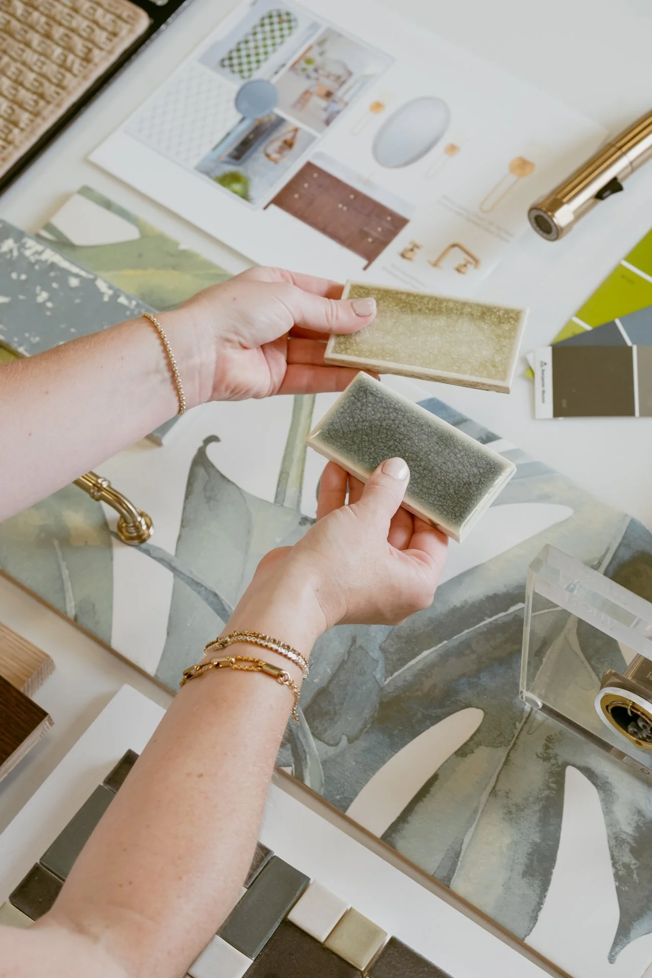 A flat lay of various tiles, finishes and a carpet sample with a women's hand holding two green tiles at the Refine Tile and Design showroom in Wilmington, North Carolina.