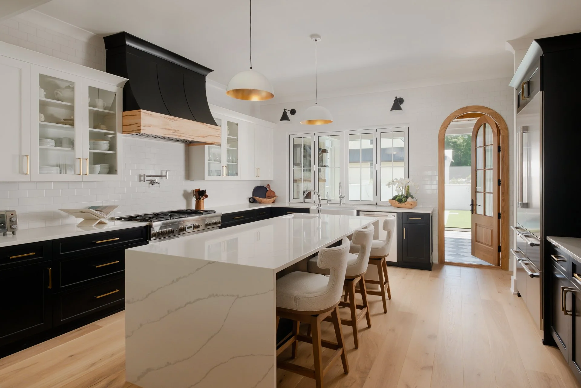 A classic kitchen with two-toned cabinets, white countertops, a waterfall island, a black range hood, wood accents, arched doorway sink. There are wood floorings, two pendant lights hanging above the island, and a full-height, all-the-way-wrapped bac