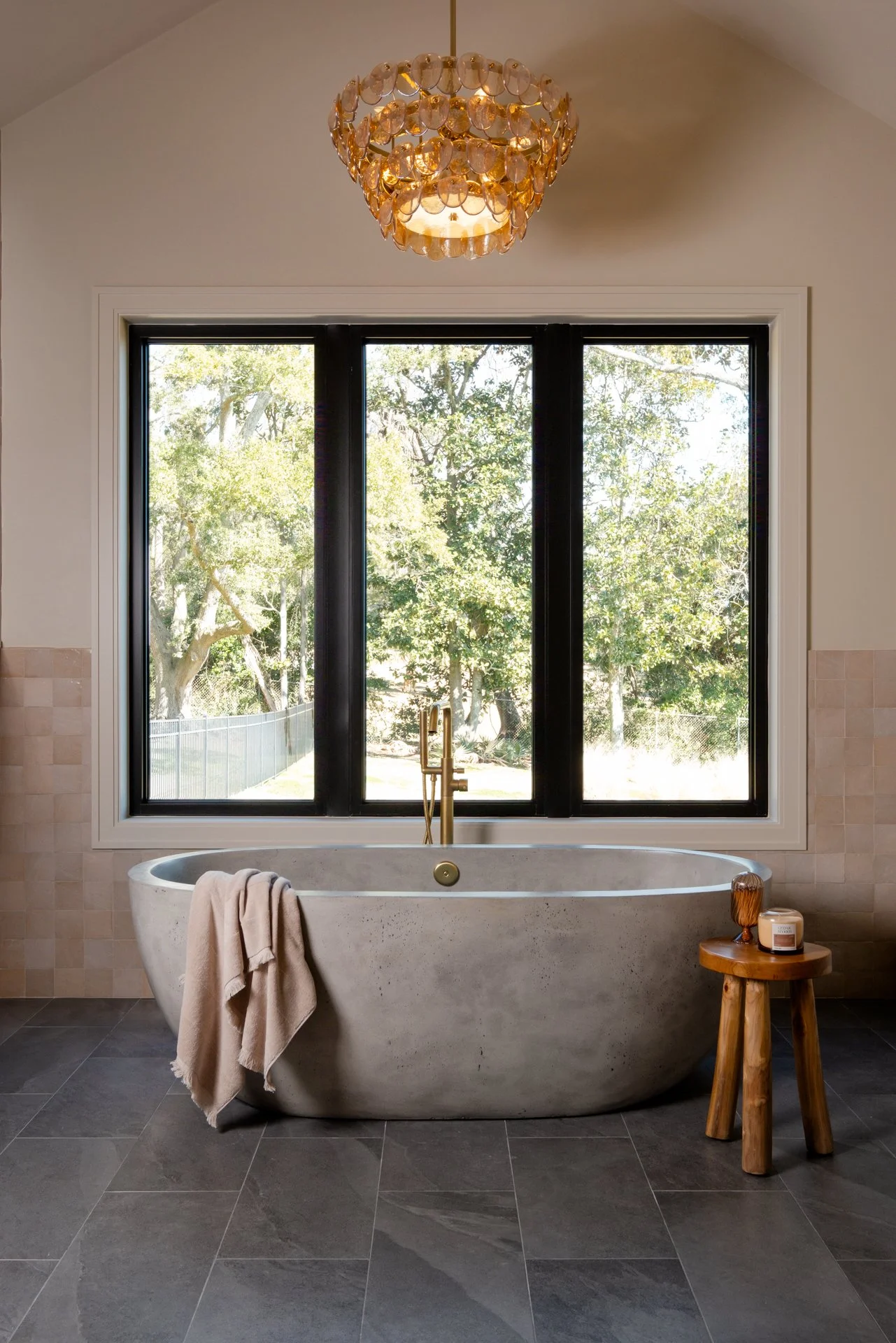 A stunning master bath concrete stand-alone soaking tub with Wayne Tile Ultra Slate Dark Gray 12x24 floor tile and a surrounding wall with 4x4 Tiles of Ezra Zellige in oat color. Large windows for natural light in the backdrop.