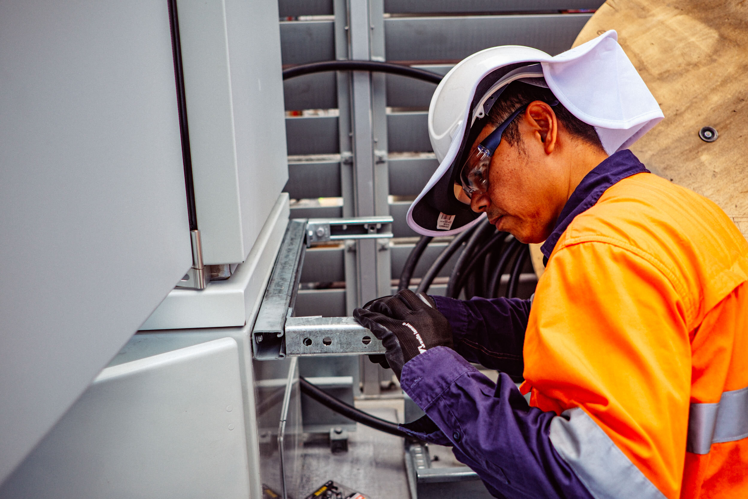 Electrician working on electrical panel, wearing a hard hat, safety glasses, and a high-visibility orange jacket, connecting wires inside a utility box.