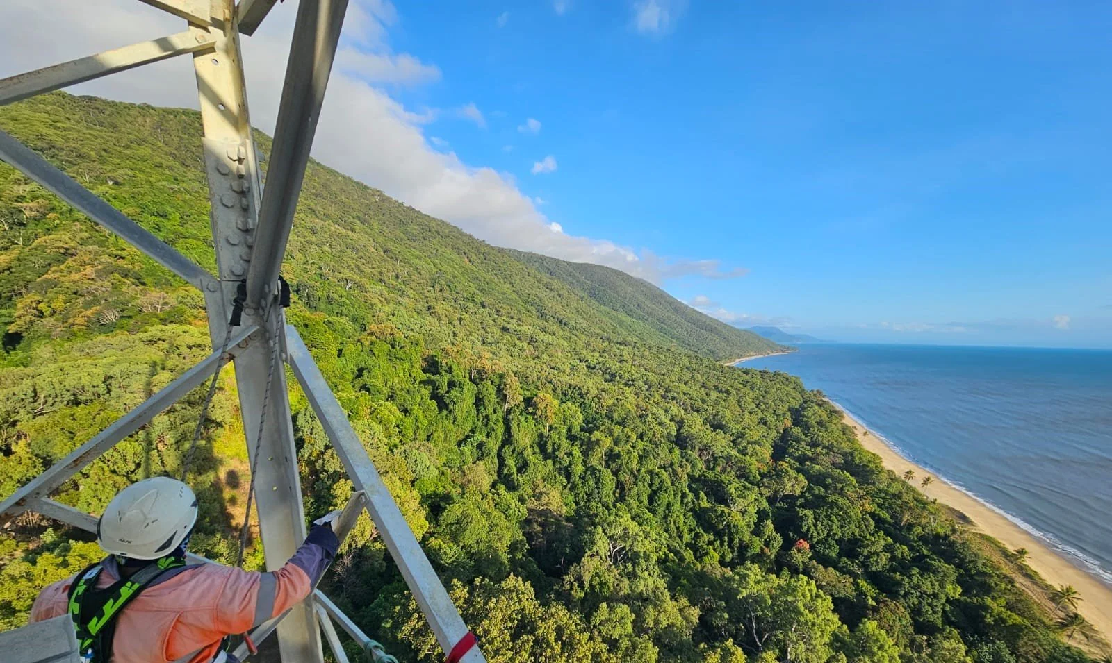 A worker in safety gear on a tall metal tower overlooking a lush green hillside and a coastline with sandy beach and calm ocean under a partly cloudy sky.