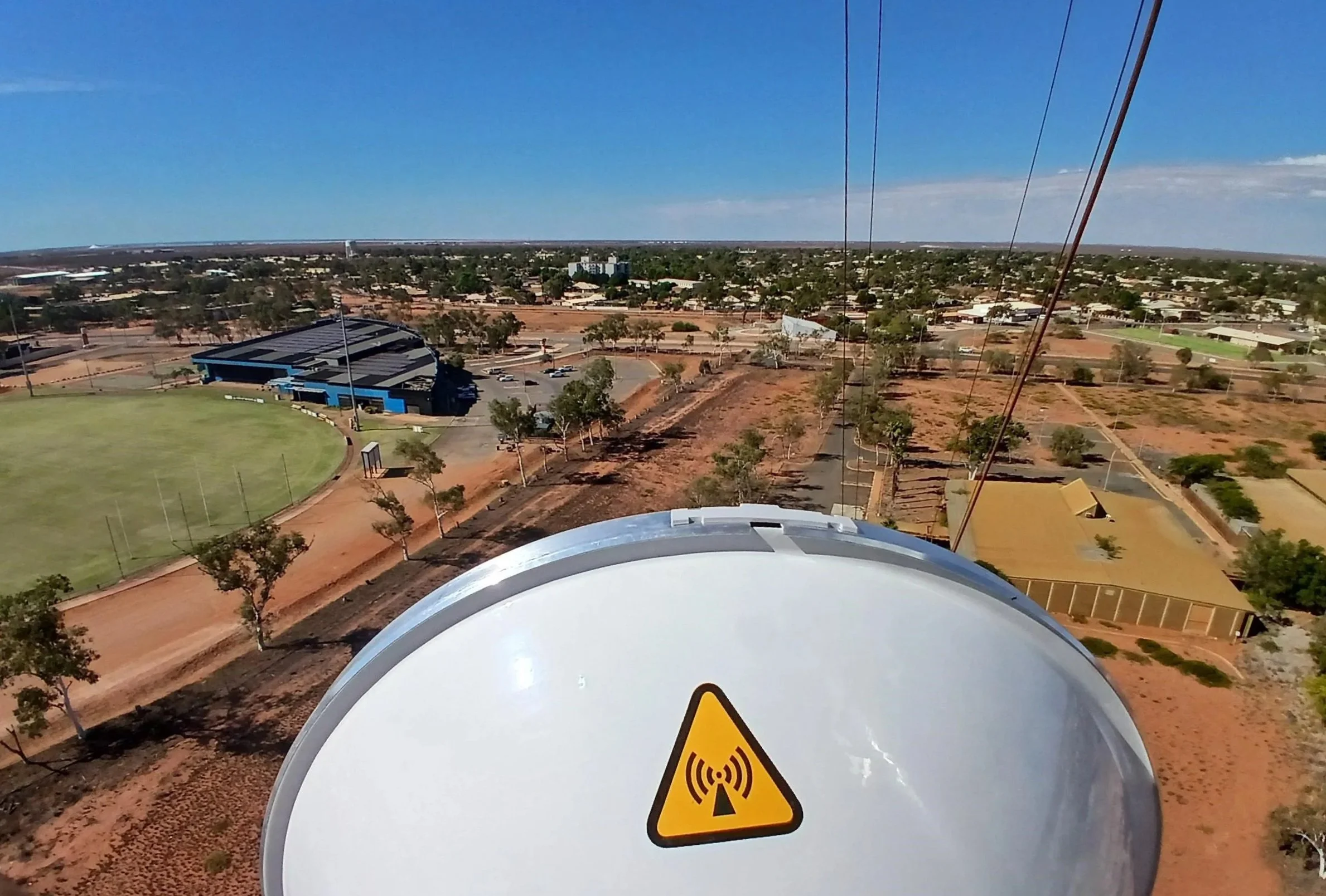 View from a tall structure showing a landscape with a sports field, buildings, trees, and dirt roads under a clear blue sky, with a large white dish with a warning sign in the foreground.